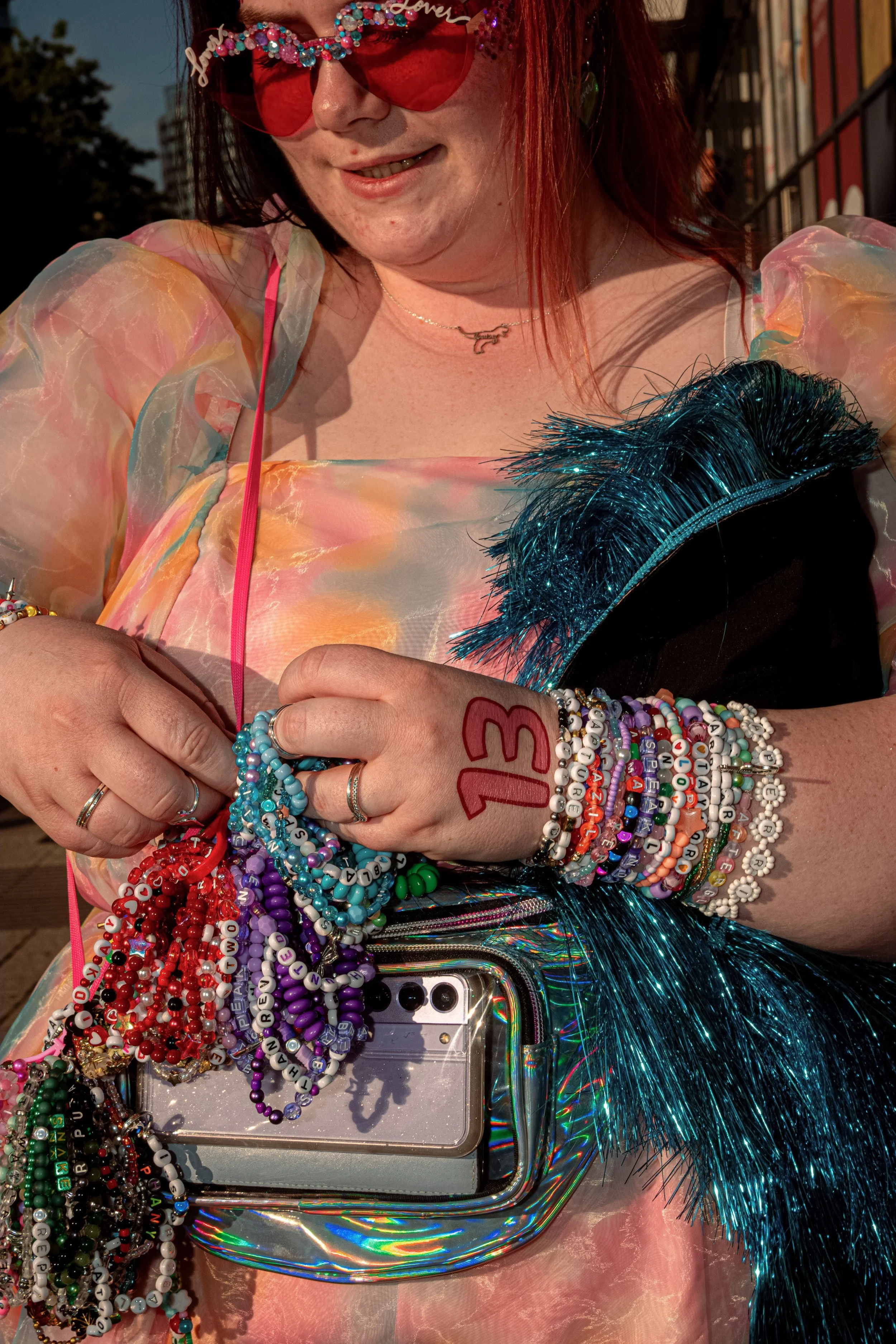 A Taylor Swift fan in red heart-shaped sunglasses adjusts friendship bracelets above a clear shoulder bag.