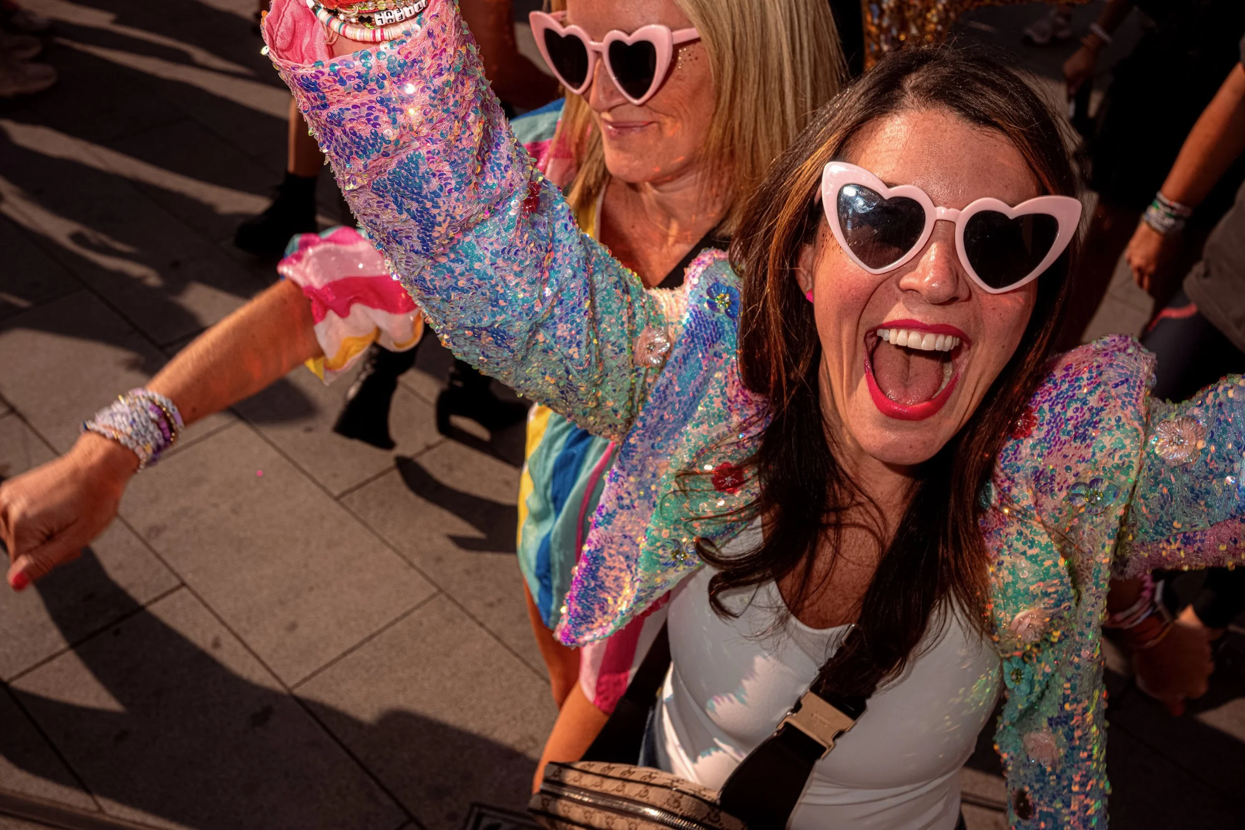 Two smiling Taylor Swift fans in sequinned jackets and pink heart-shaped sunglasses raise an arm in the crowd.