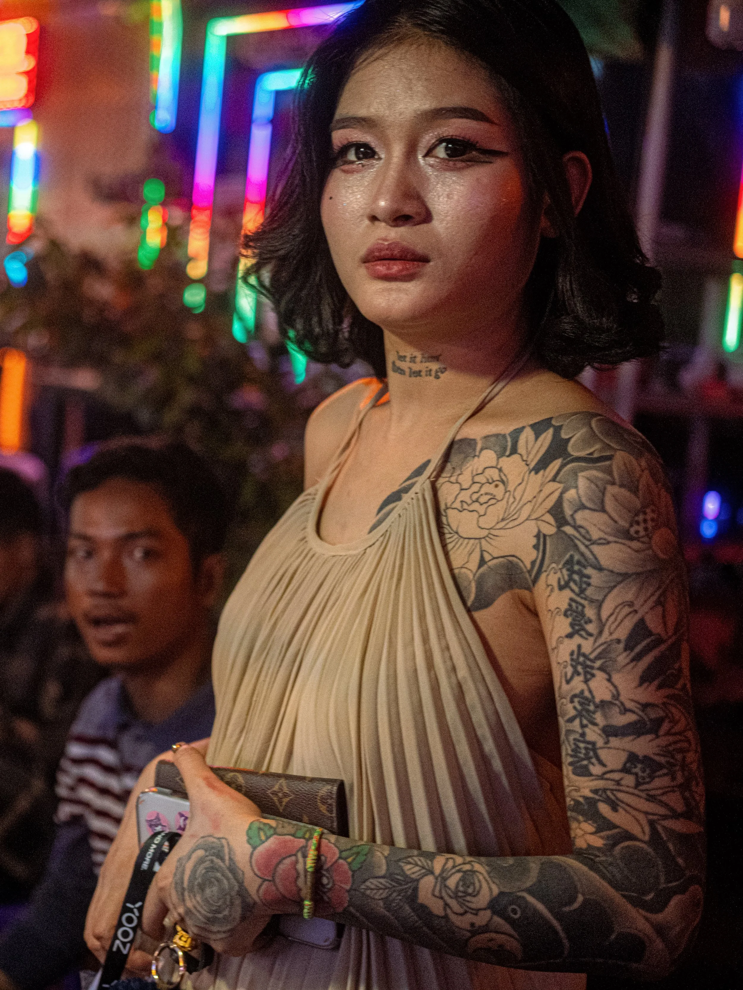 A tattooed person in a beige pleated outfit stands beside a seated person in a neon-lit bar in Thailand.