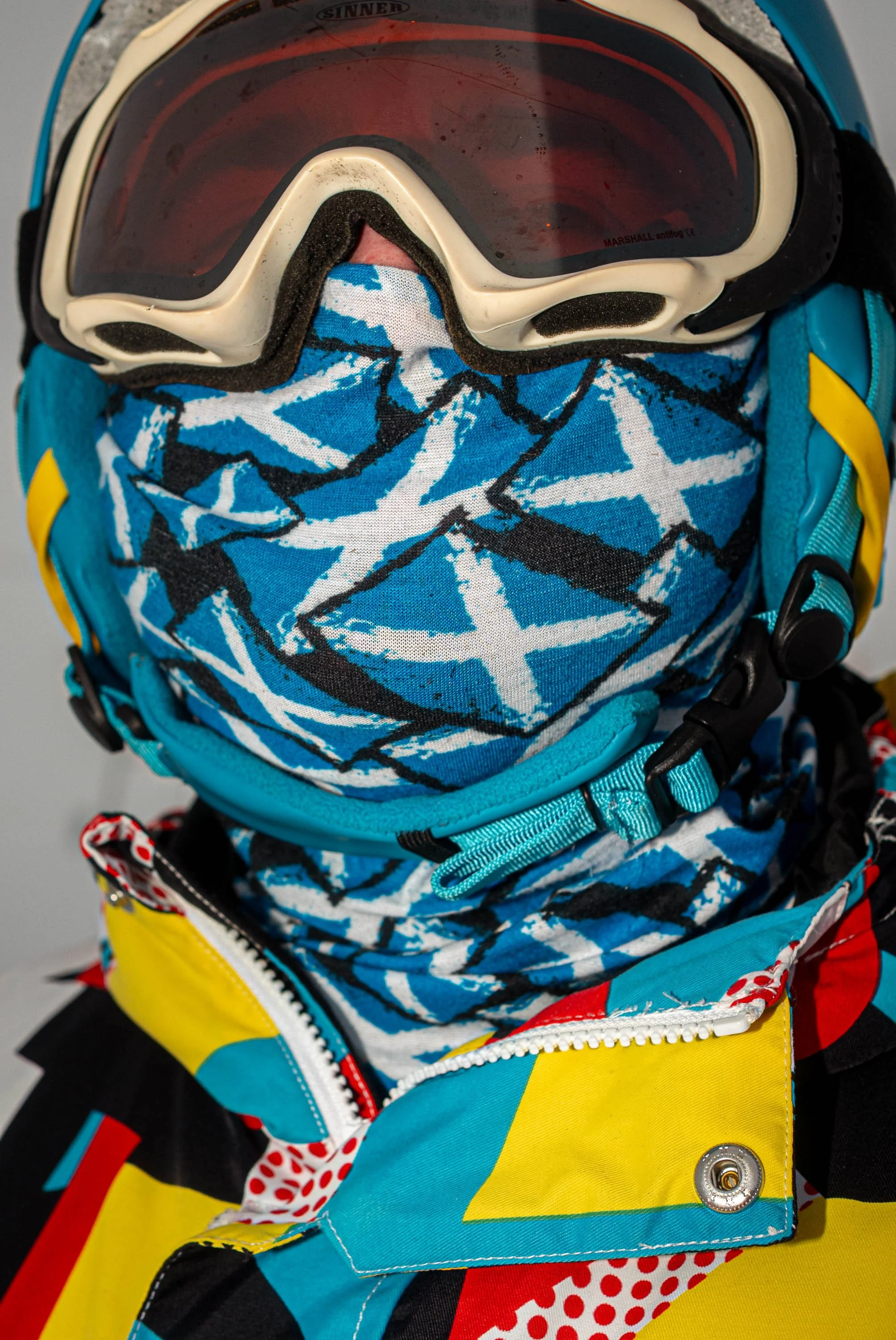 Close-up of a skier hidden behind goggles and a Saltire-print snood, above a bright retro ski jacket.