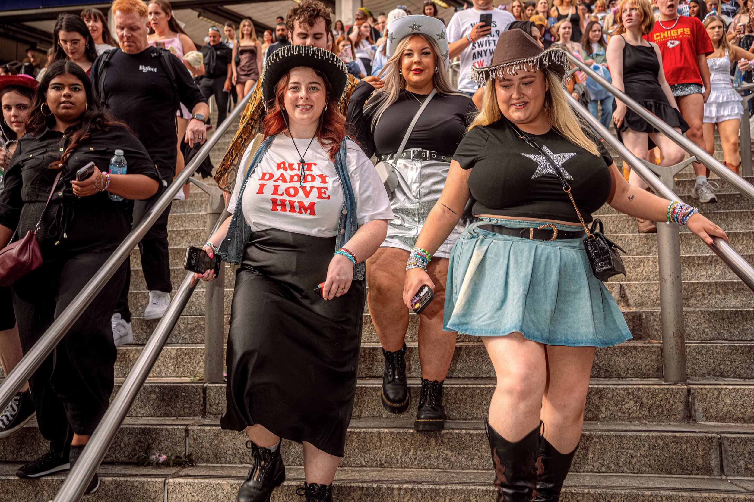 Taylor Swift fans descend a crowded staircase wearing cowboy hats, boots and concert outfits.