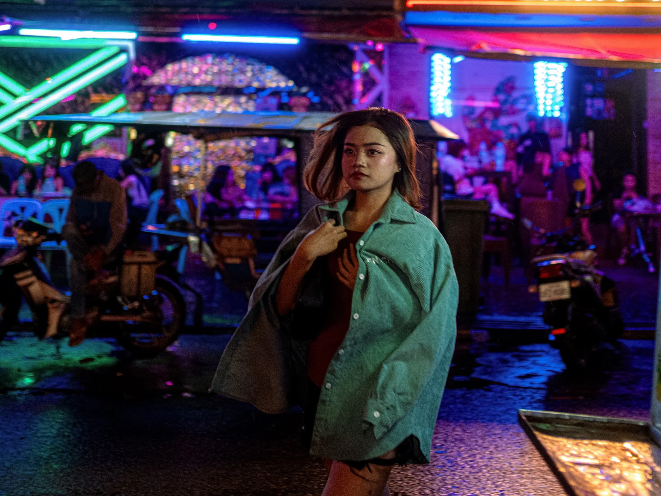 A person with a light jacket pulled over their head stands outside a neon-lit bar.