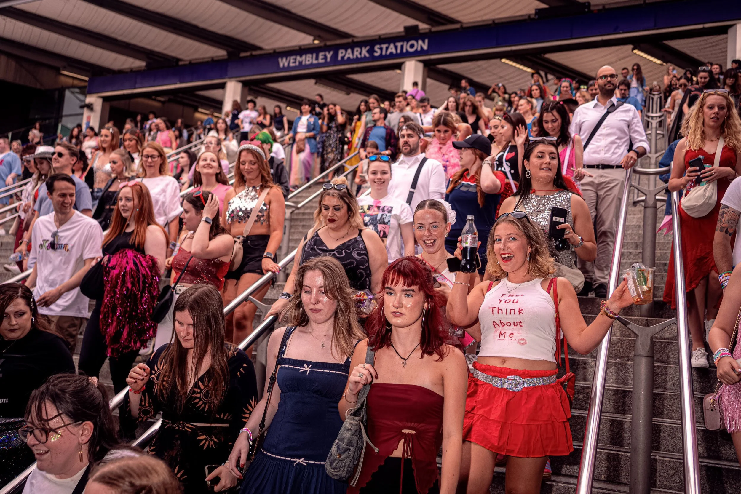 A crowd of Taylor Swift fans in colourful concert outfits descends the steps at Wembley Park station.