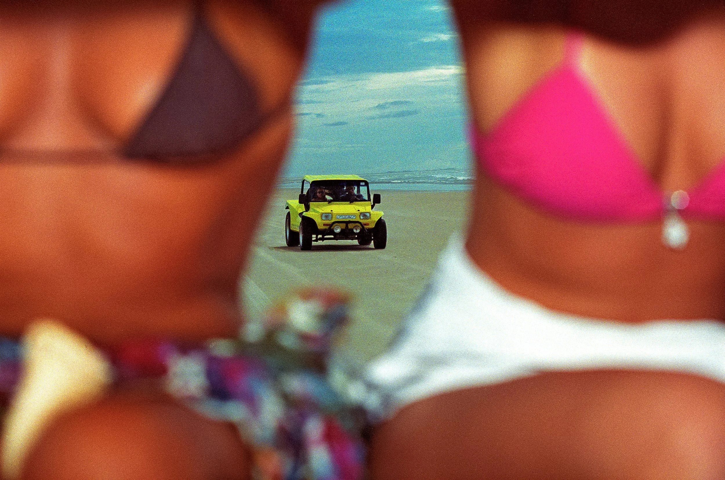 A yellow beach buggy is framed between two seated holidaymakers in bikinis.