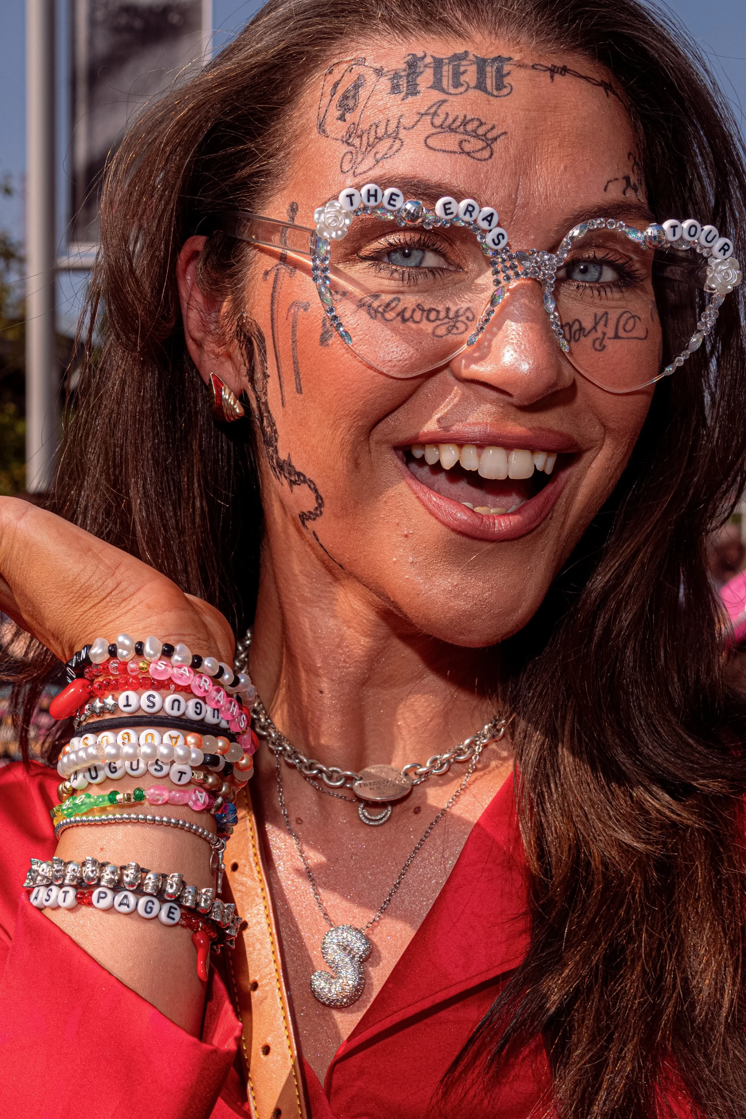 A smiling Taylor Swift fan wears clear-framed glasses, lettering painted across her face and stacked friendship bracelets.