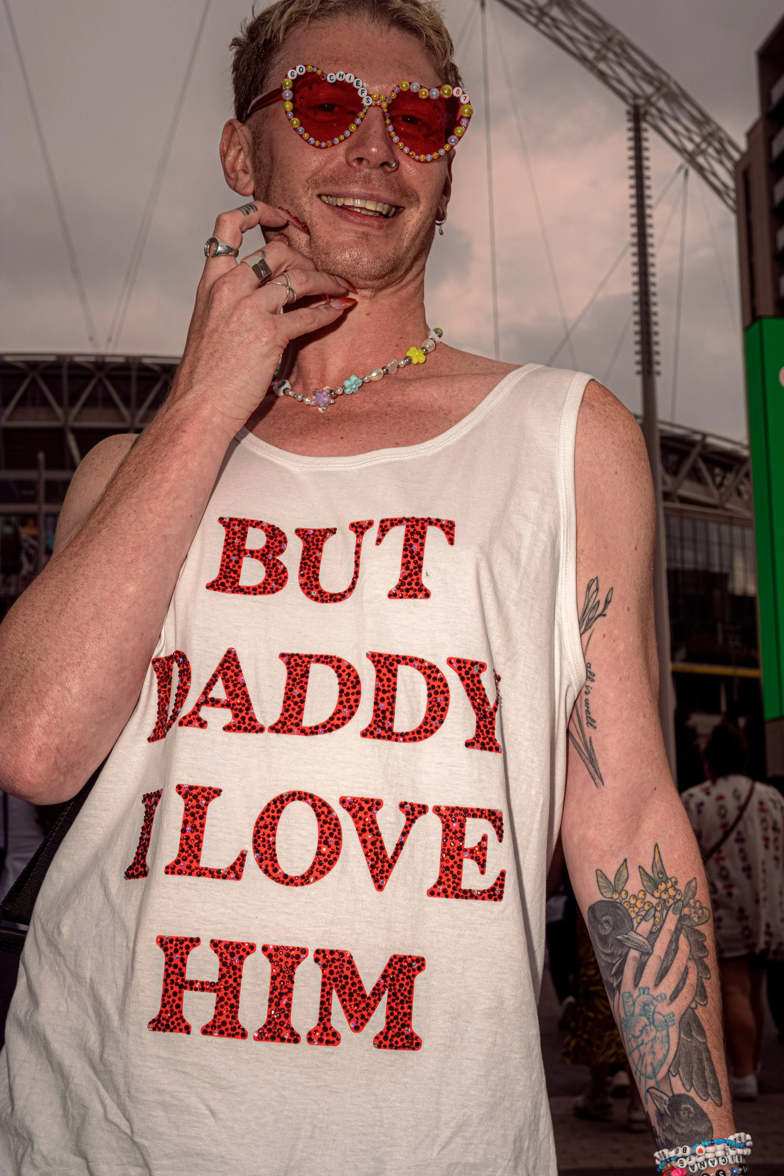 A smiling Taylor Swift fan wears red heart-shaped sunglasses and a white vest reading “But Daddy I Love Him”.