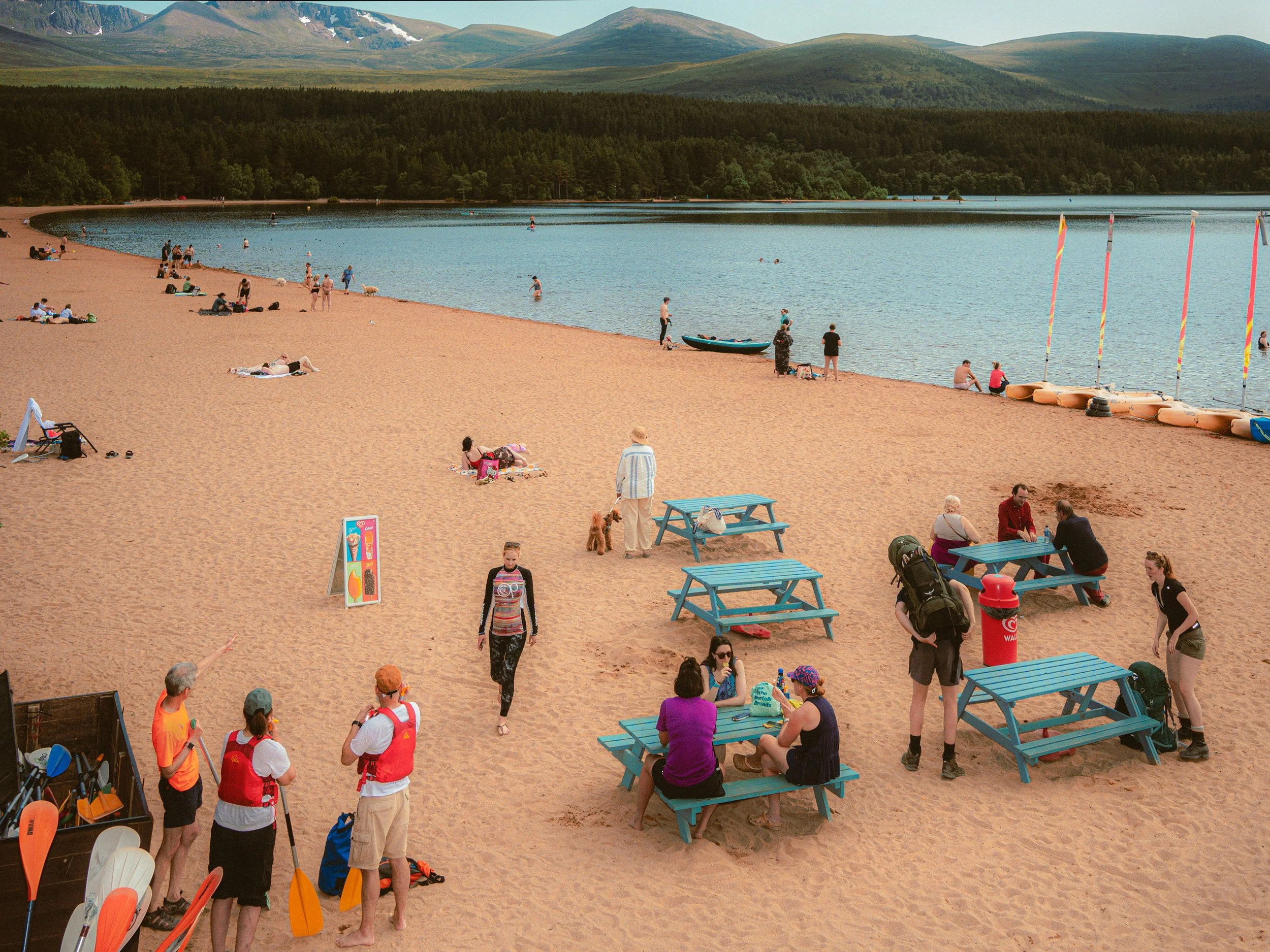 Visitors relax on a sandy loch beach with picnic tables and mountains behind.