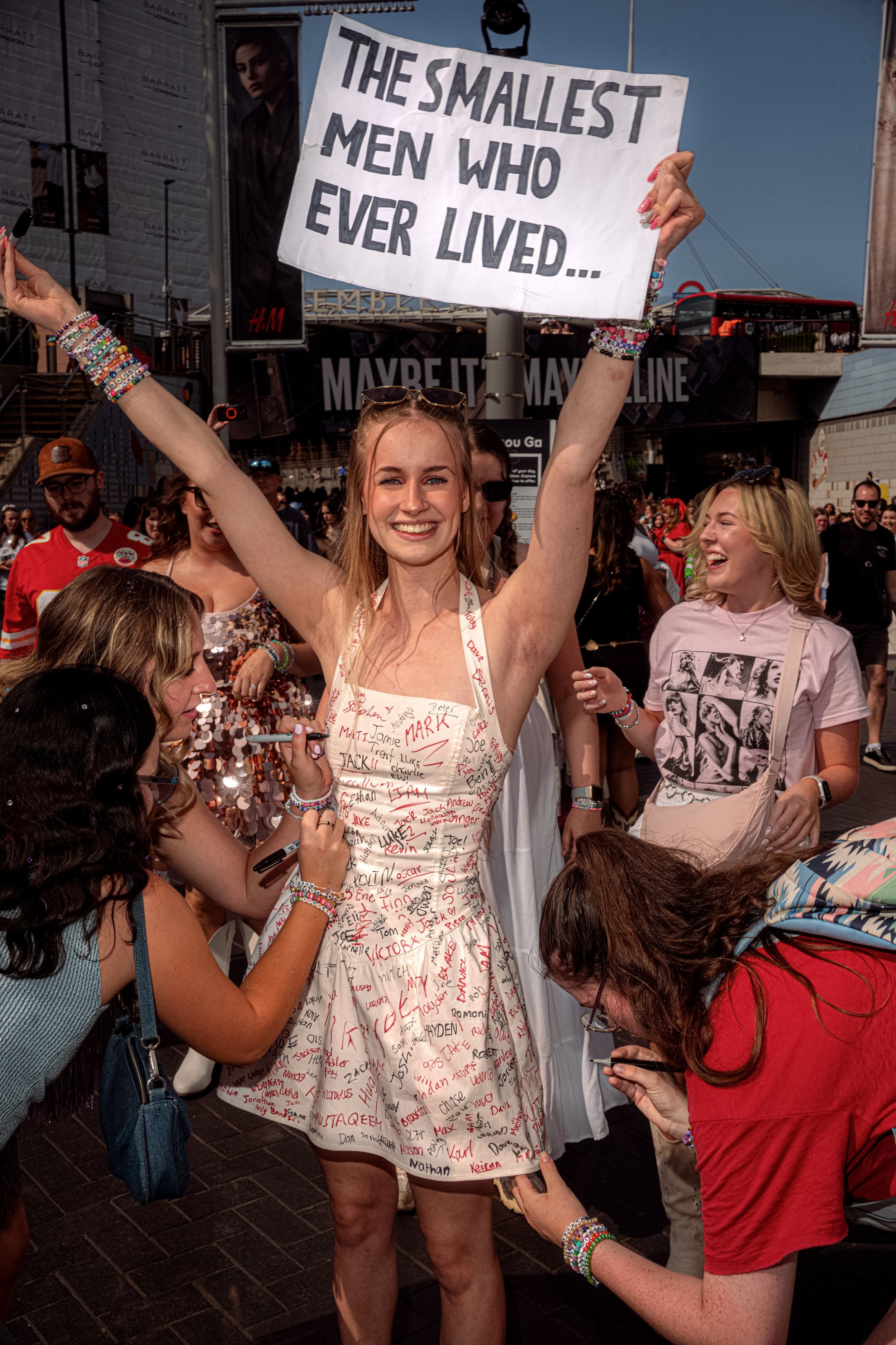 A Taylor Swift fan in a silver dress raises a sign reading “The smallest men who ever lived” above a group of fans.
