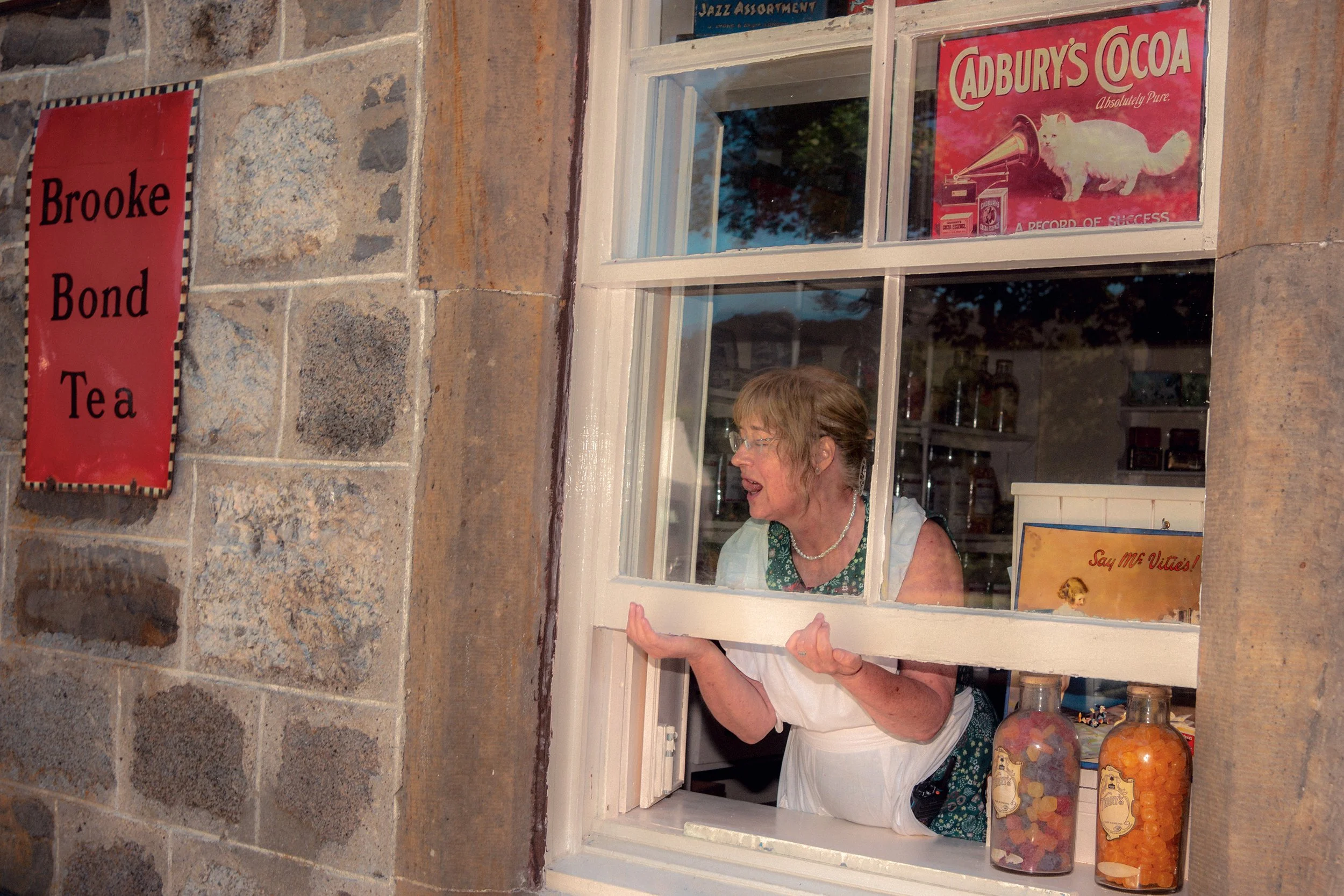 A woman leans out of the window of a stone shop with old Brooke Bond Tea and Cadbury's Cocoa signs.