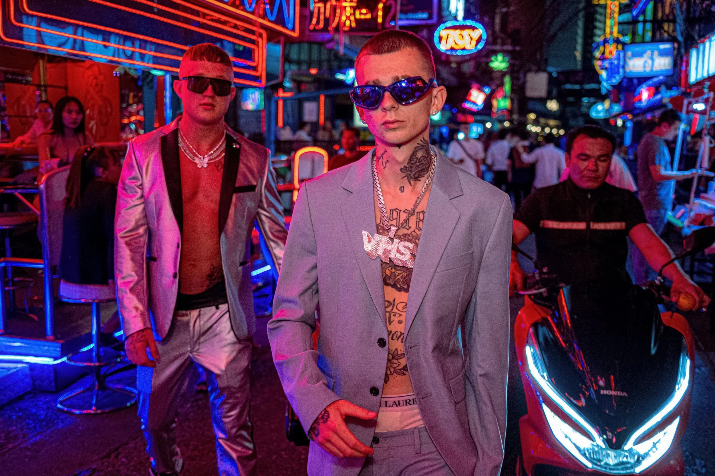 Two people in pastel suits and sunglasses walk along a neon-lit street beside a motorbike in Thailand.
