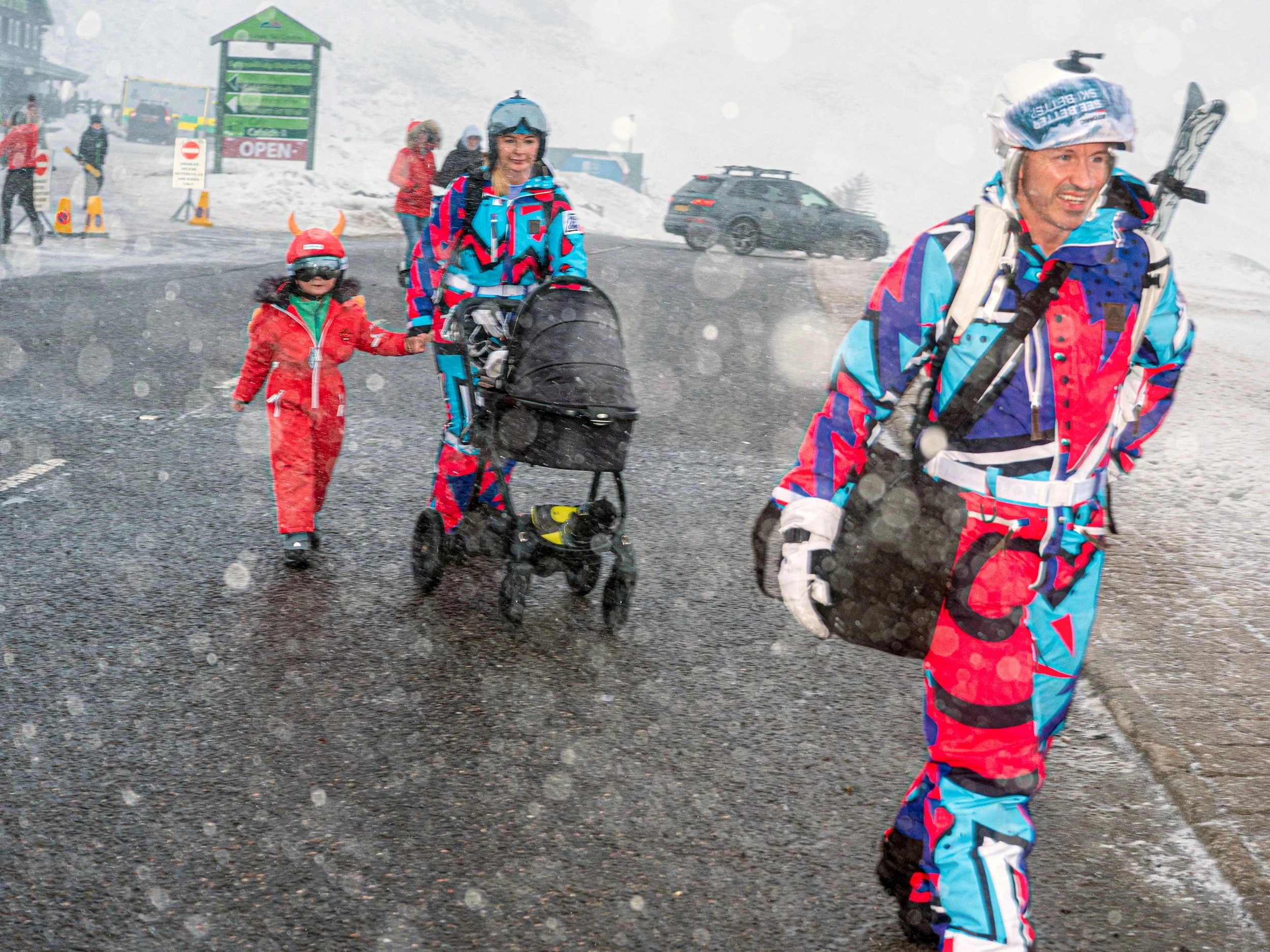 A skier in a bright retro one-piece suit walks through falling snow with a child and a pushchair.