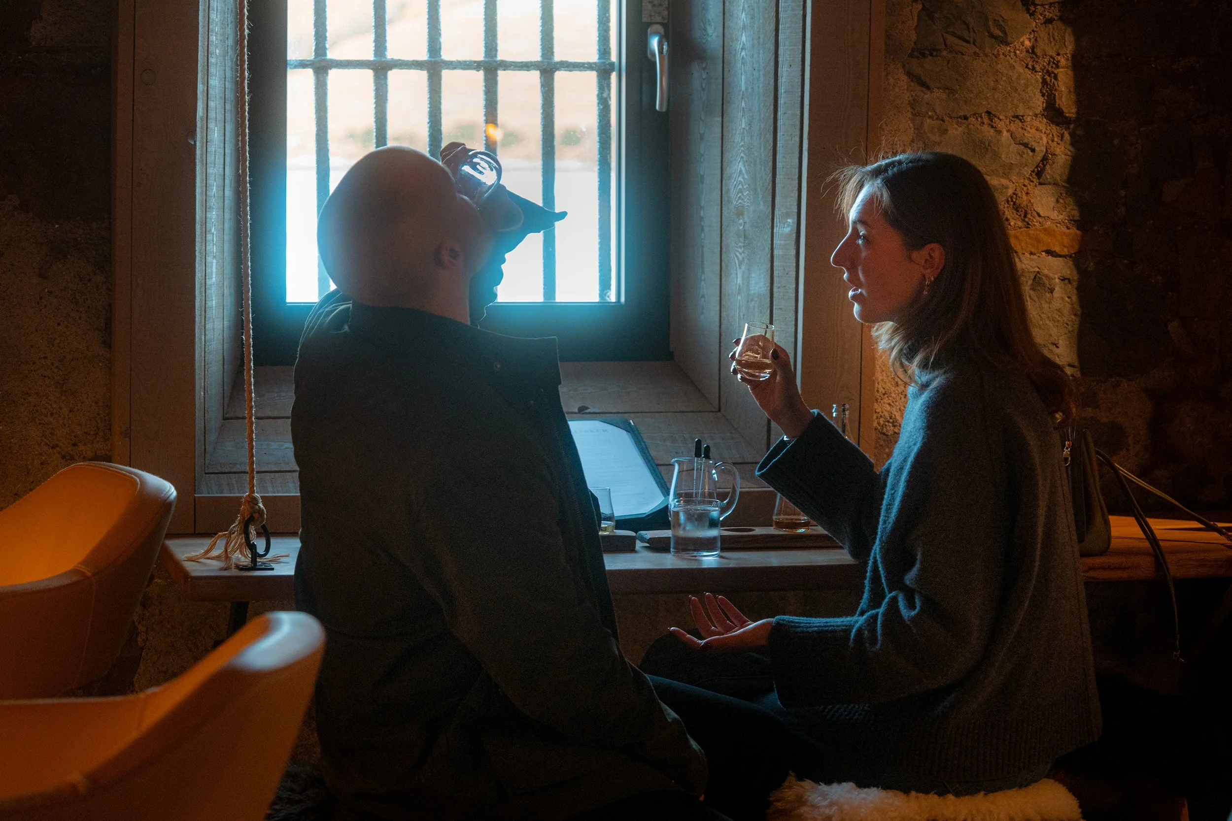 Two people sit at a table by a barred window in a stone-walled room, one with a laptop.