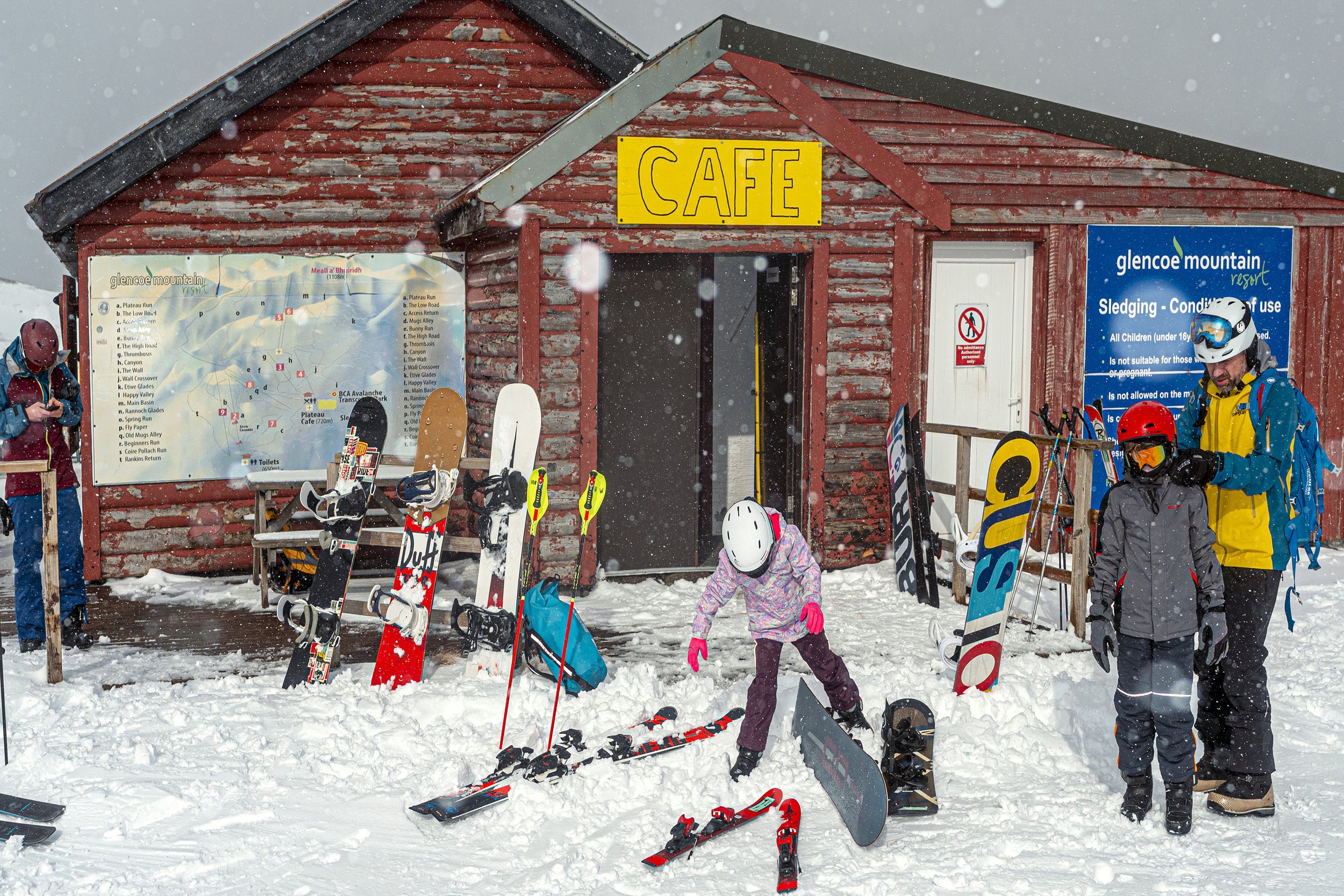 Skiers and snowboarders sort their kit outside a red wooden café at Glencoe Mountain in the snow.