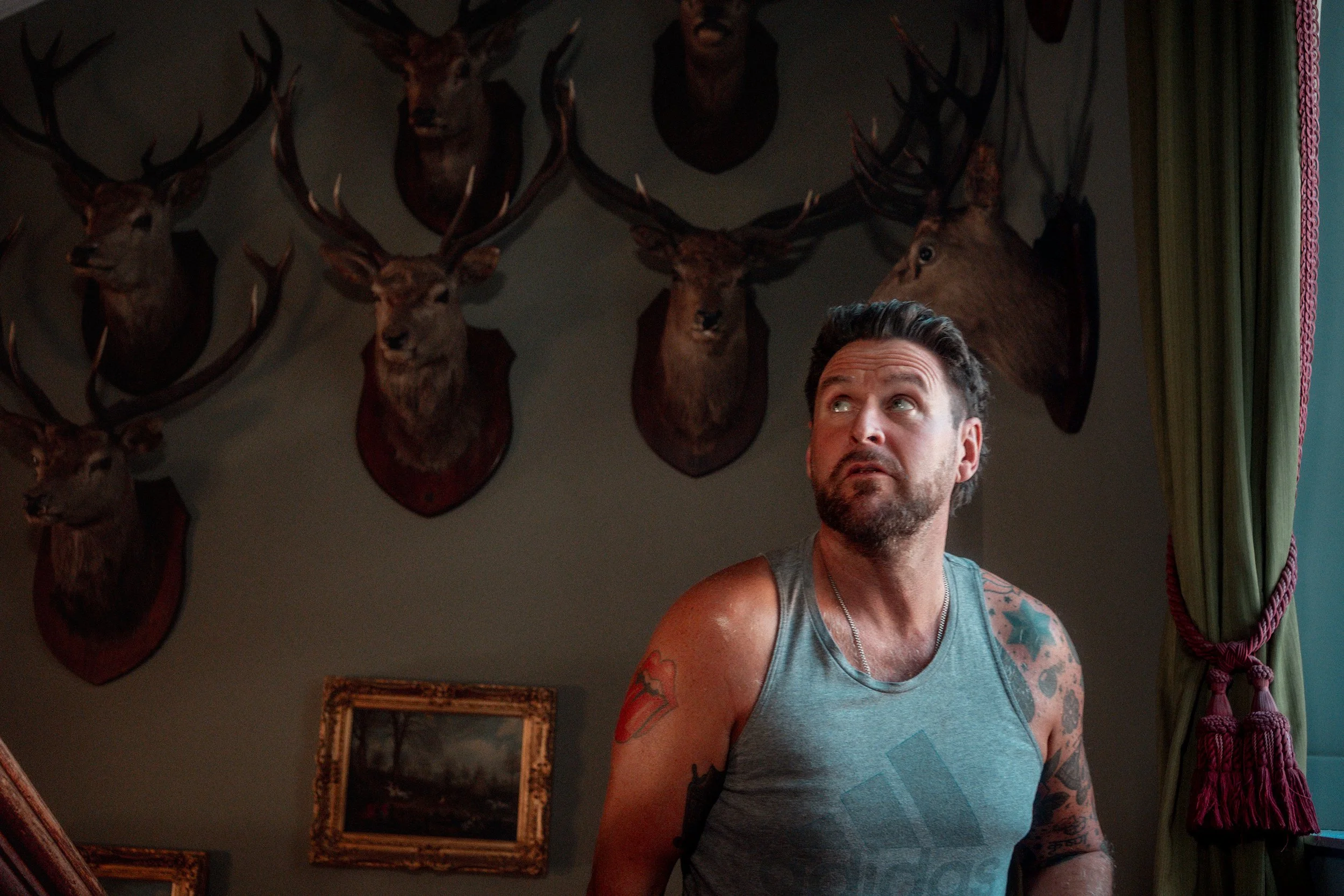 A tattooed man in a vest stands beneath mounted stag heads in a castle room.