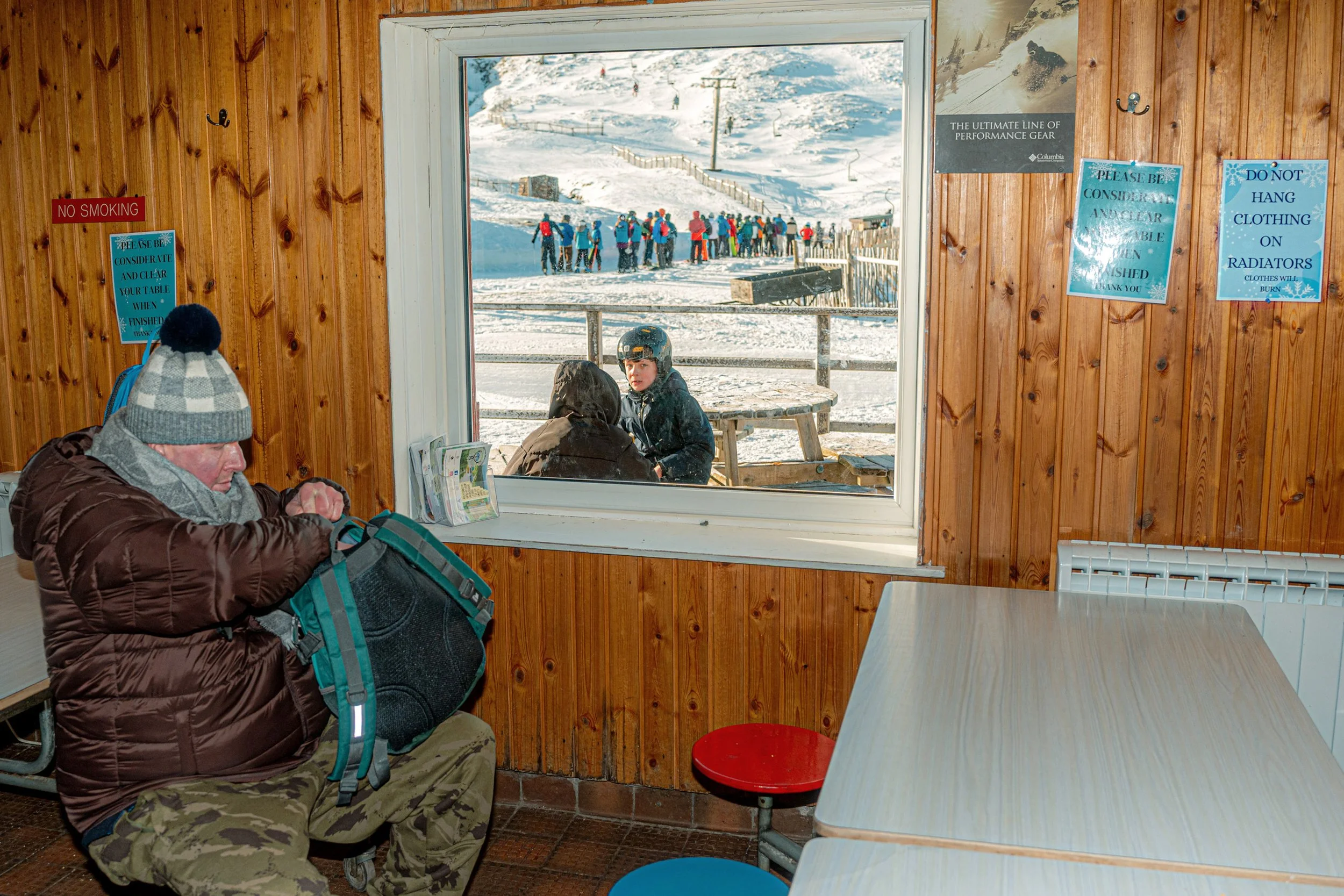 A man in a bobble hat sits in a wood-panelled café while skiers gather outside the window.