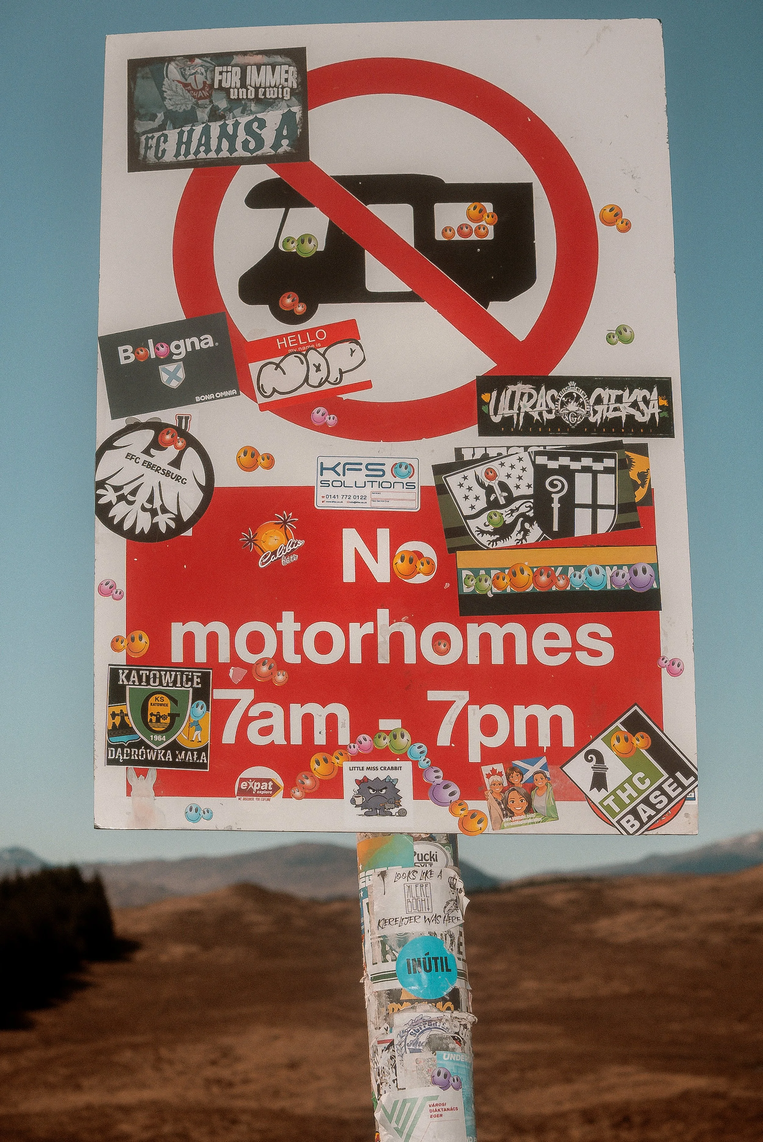 A No Motorhomes 7am to 7pm sign covered in stickers stands against the hills.