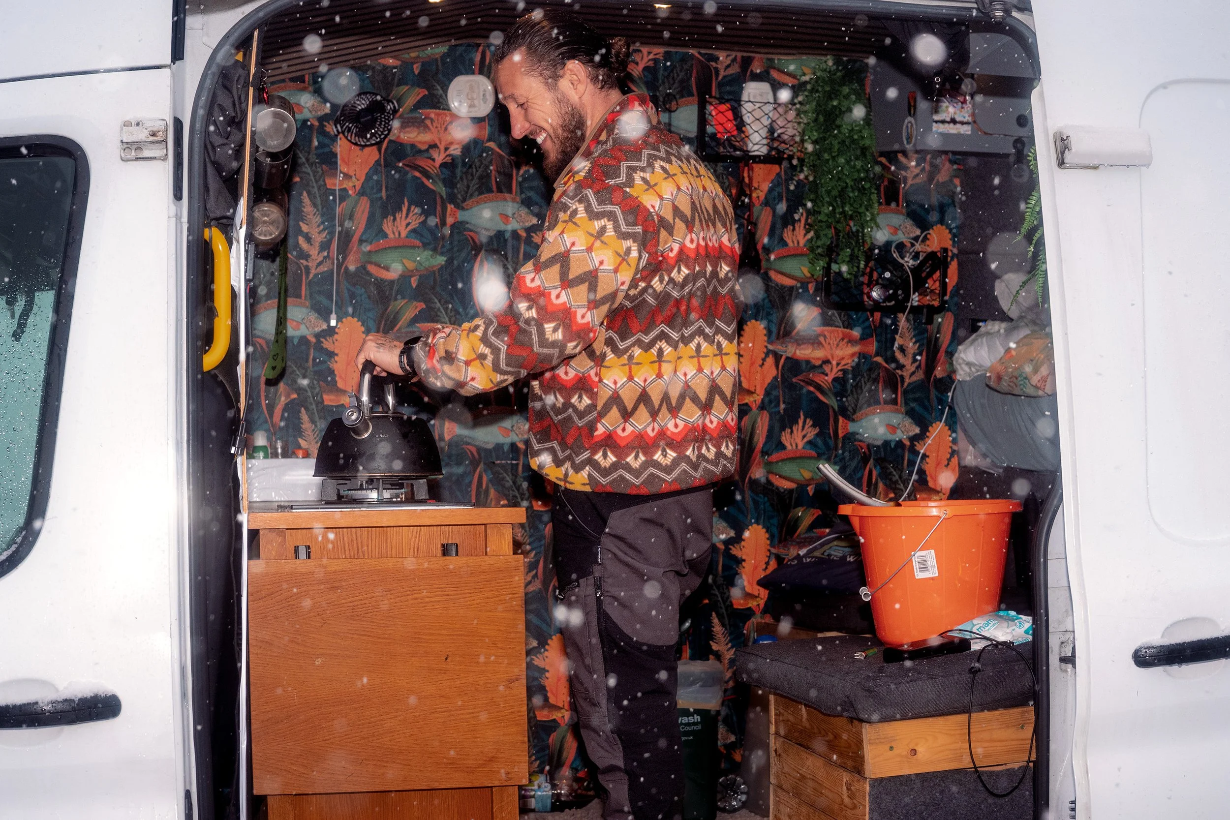 A man in a patterned jumper makes a hot drink in the back of his camper van as snow falls.