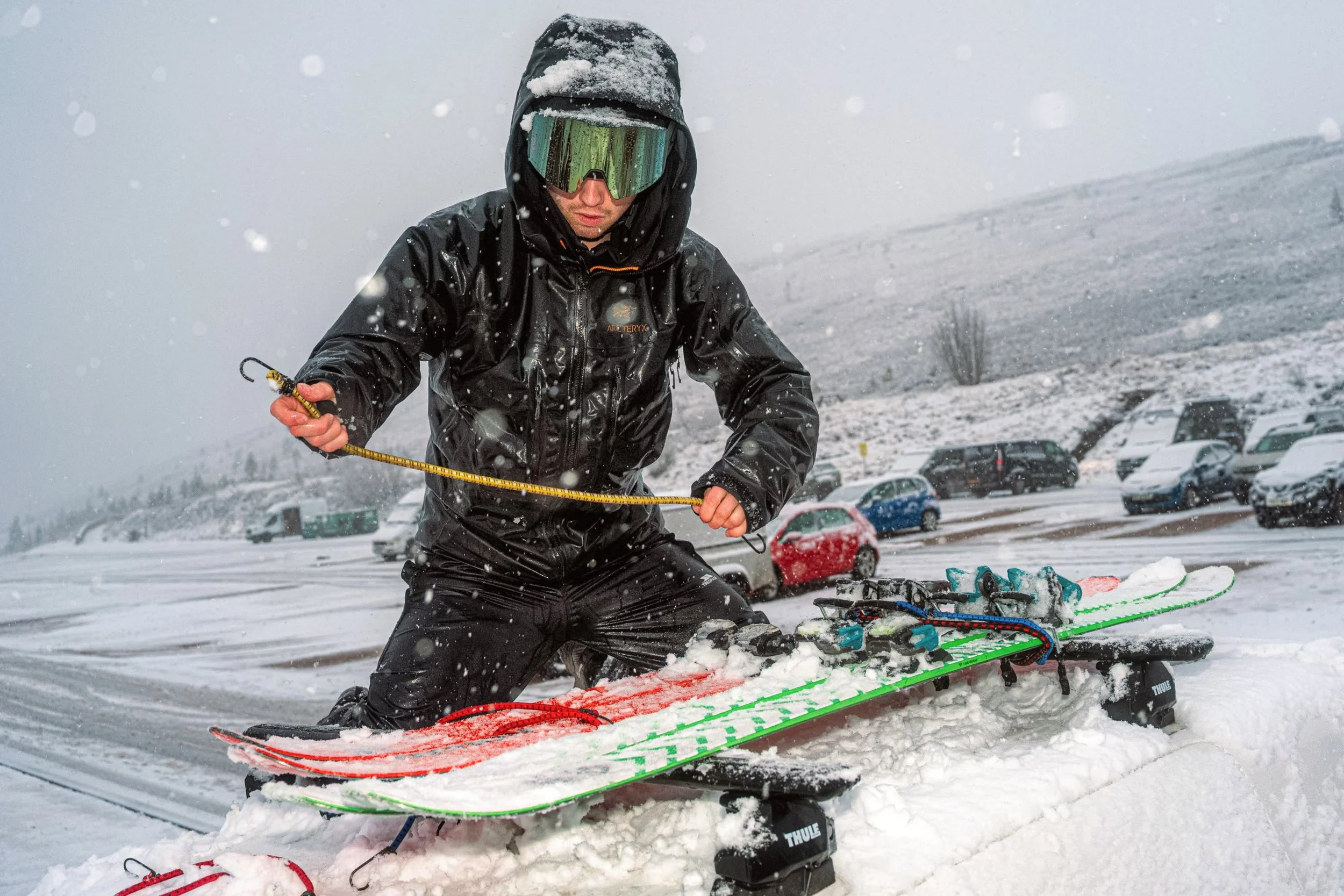 A skier in dark clothing and goggles handles equipment on a snowy car roof, from Weather Permitting.