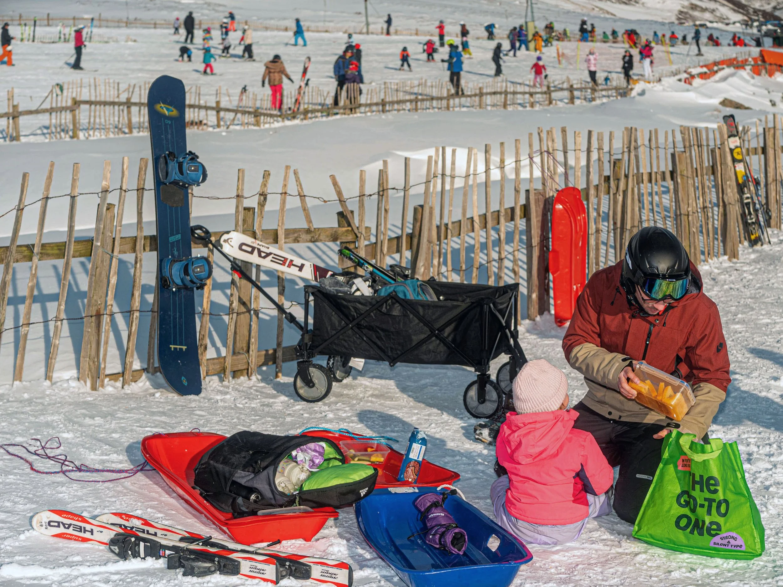 A man and a child sort through sledges and bags beside a fence at a busy ski area.