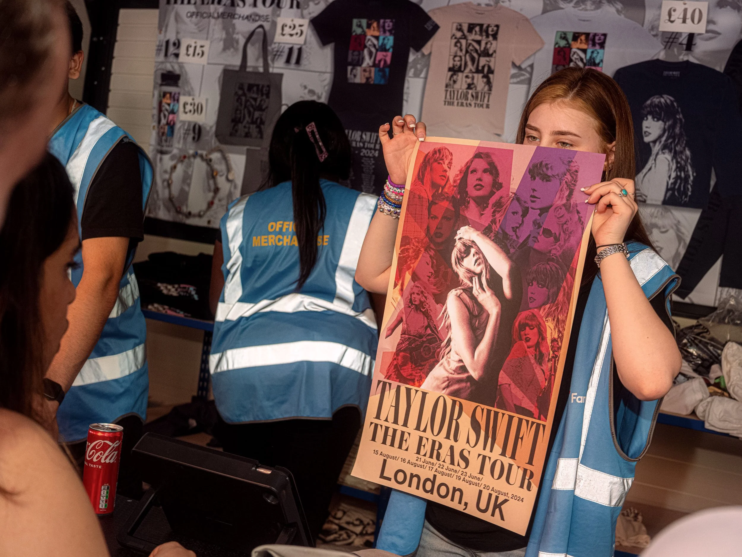A merchandise seller holds up a Taylor Swift Eras Tour London poster in front of a stall of T-shirts.