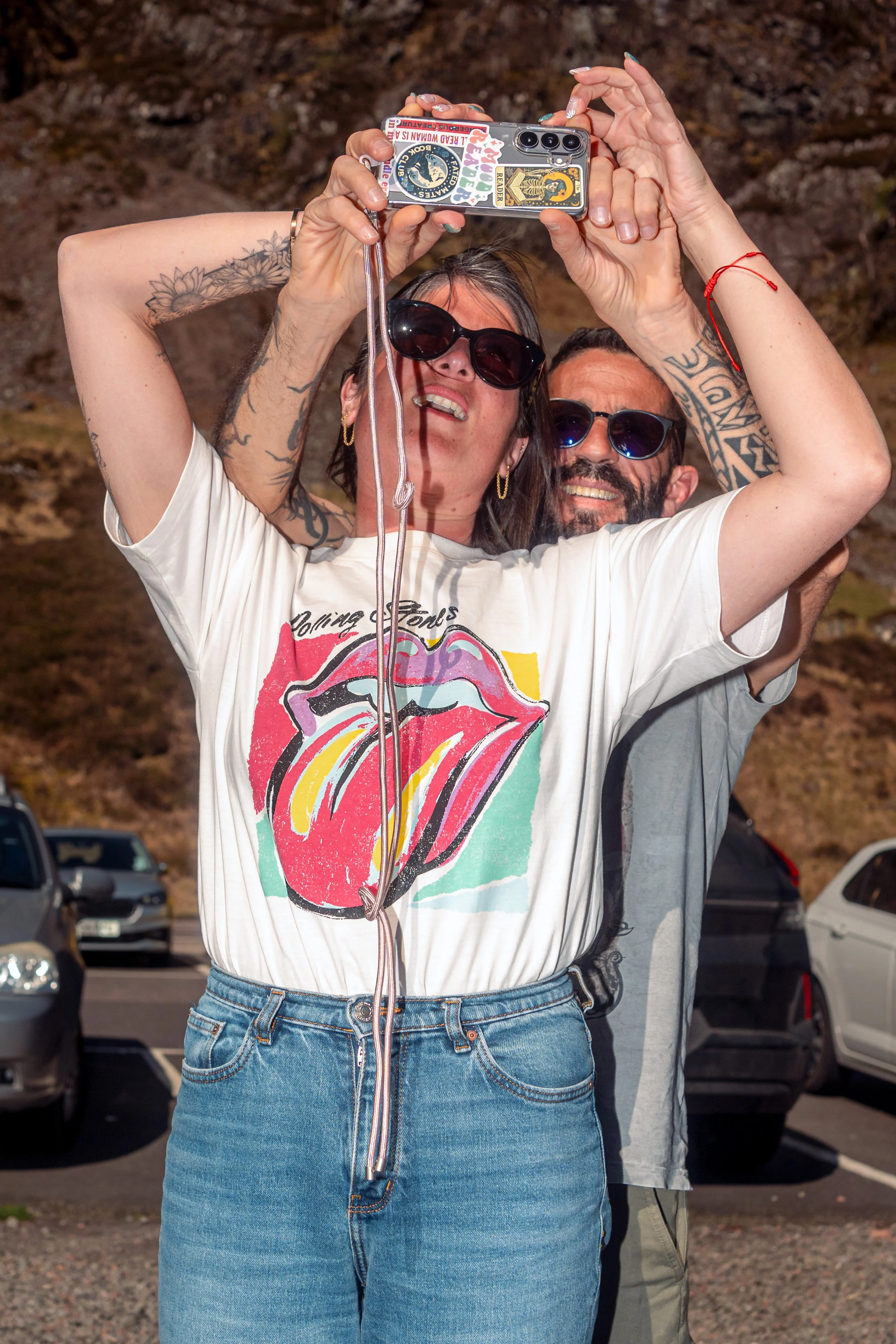 A couple in sunglasses take a selfie with a camera held above their heads in a car park.