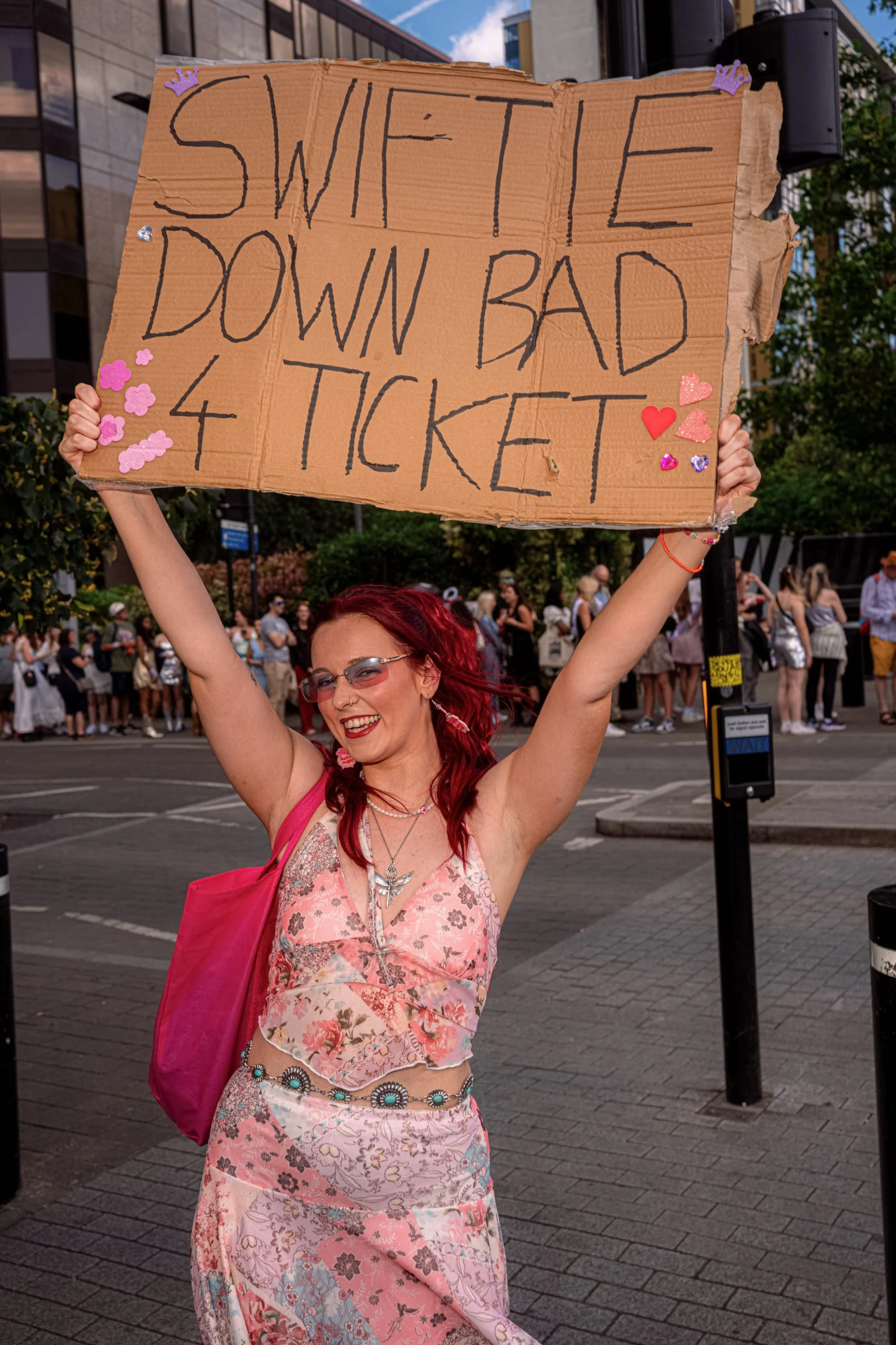 A Taylor Swift fan raises a cardboard sign reading “Swiftie down bad 4 ticket” above her head.