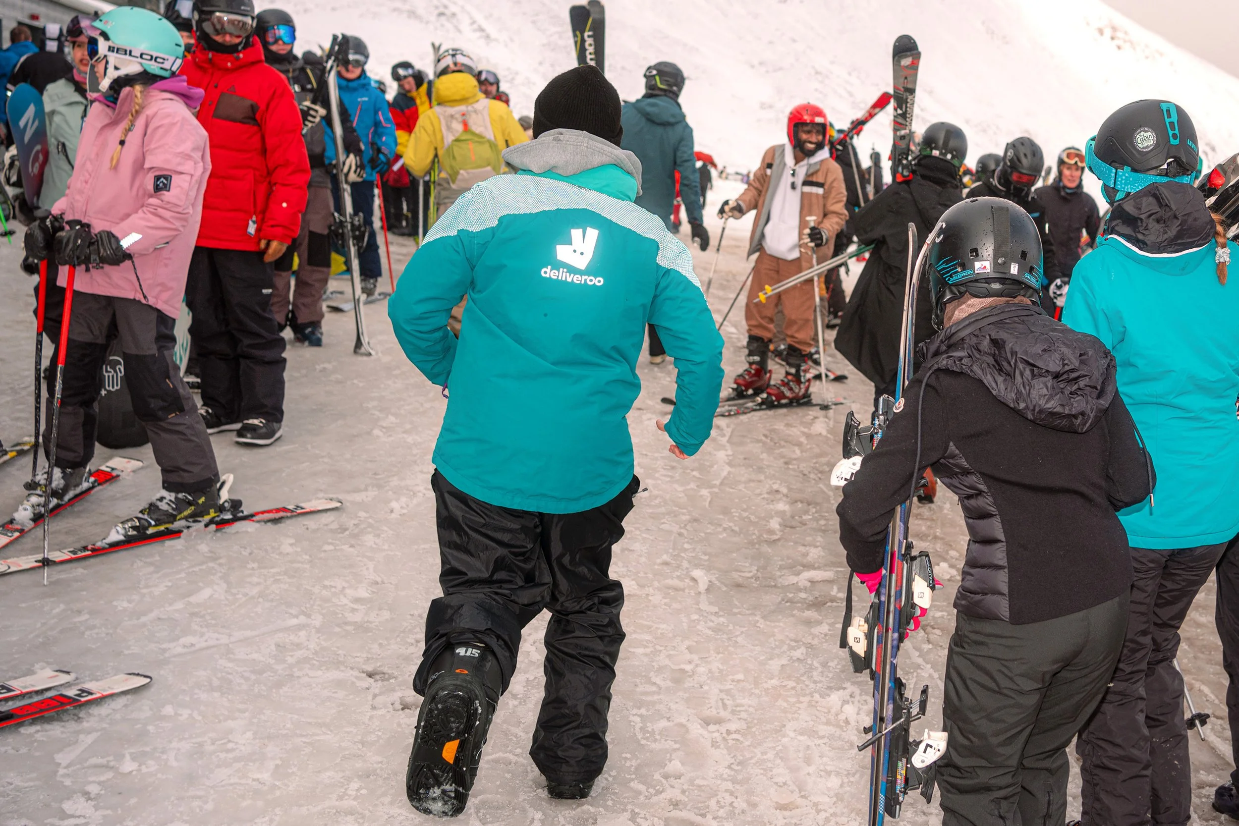 Skiers and snowboarders in colourful jackets queue at a ski centre, one in a Deliveroo jacket.