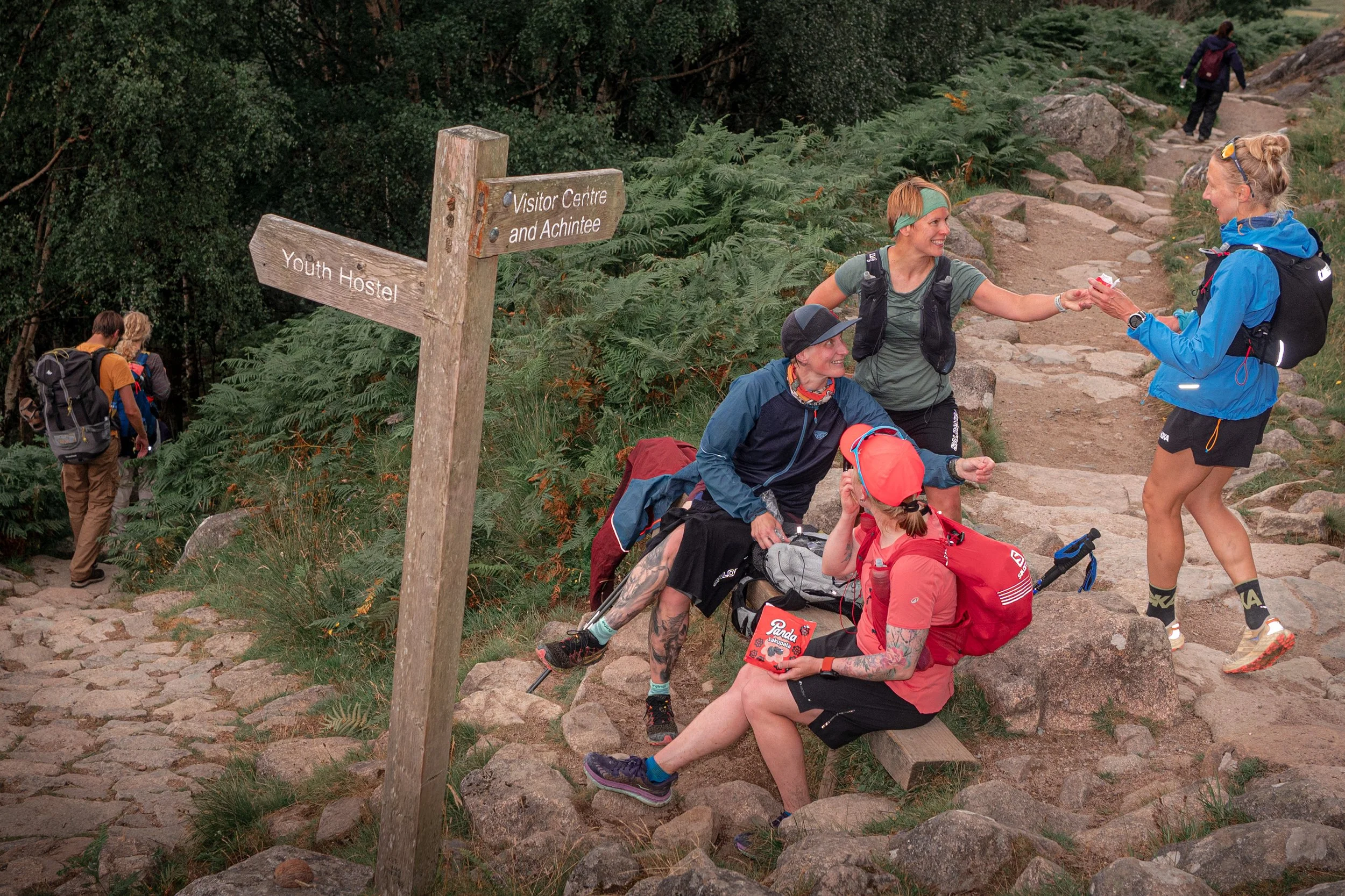Hikers rest and high-five on a stone path beside a wooden signpost.