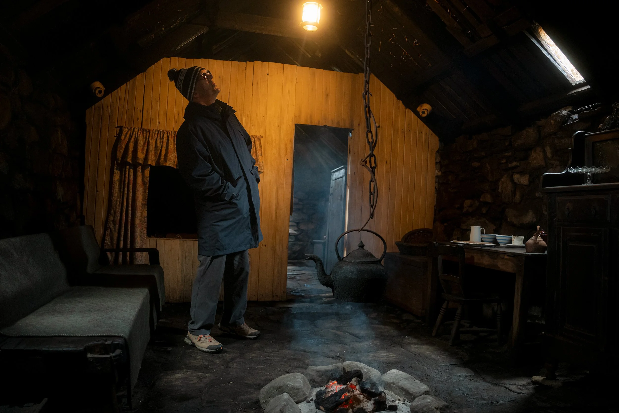 A visitor stands in the dim, smoky interior of a traditional blackhouse with a peat fire.