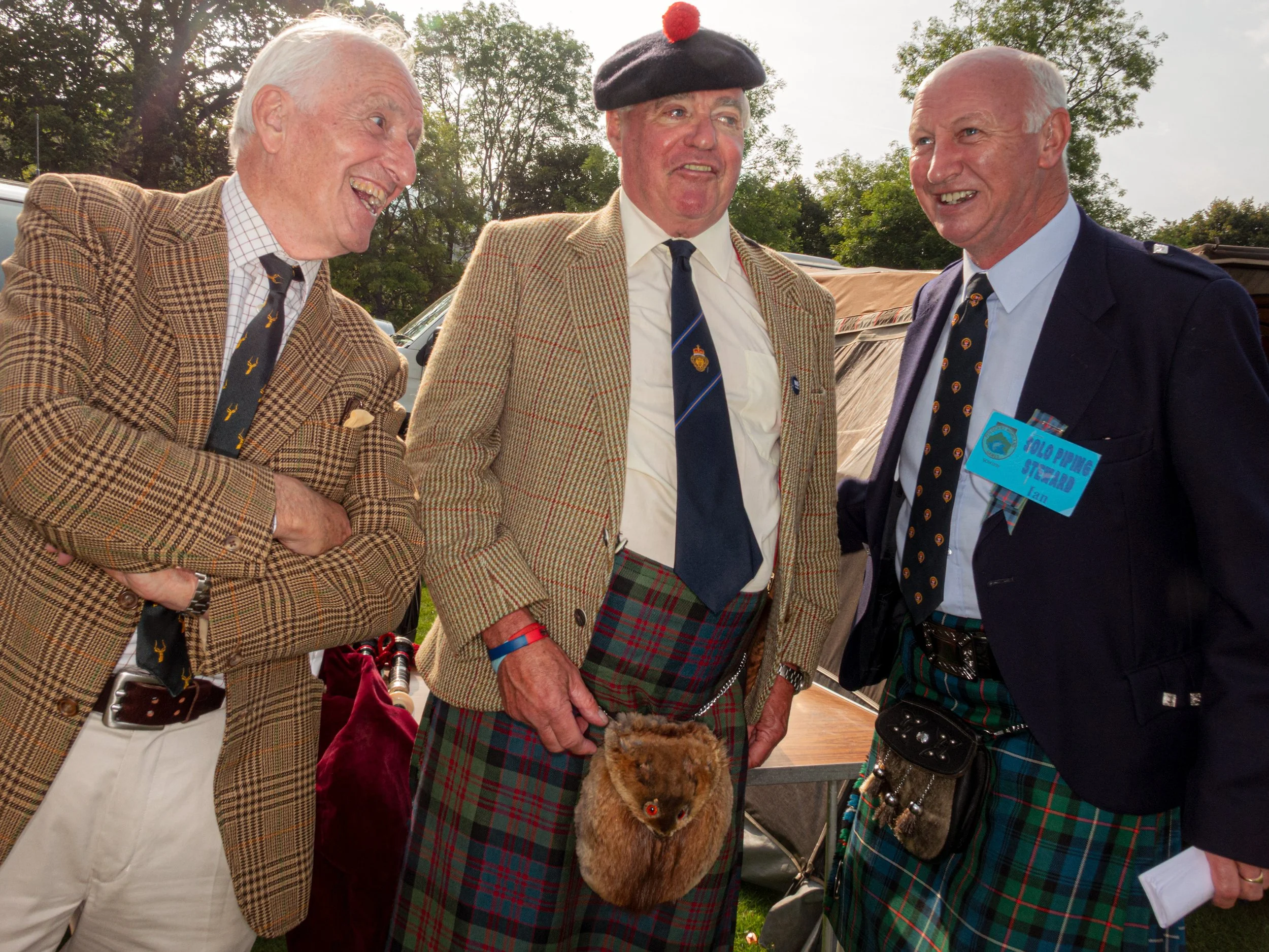 Three older men in tweed jackets and kilts laugh together at a Highland gathering.