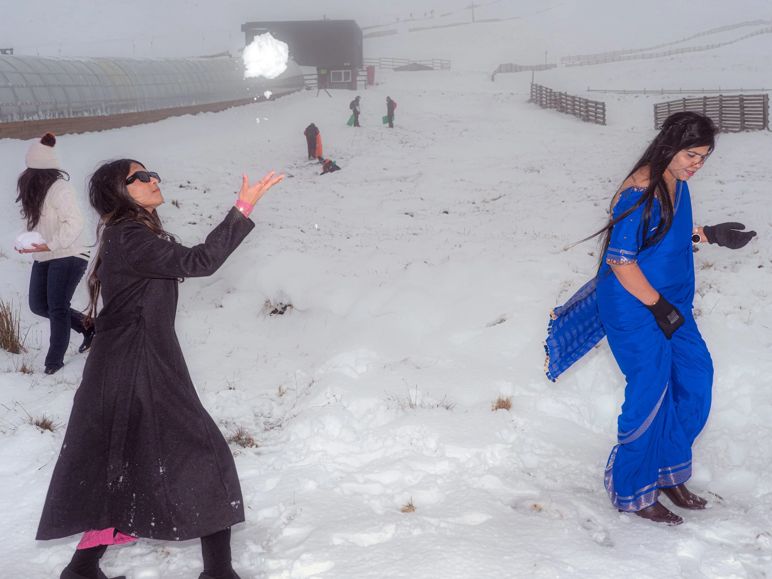 Two women, one in a long black coat and one in a blue sari, throw snow on a misty slope.