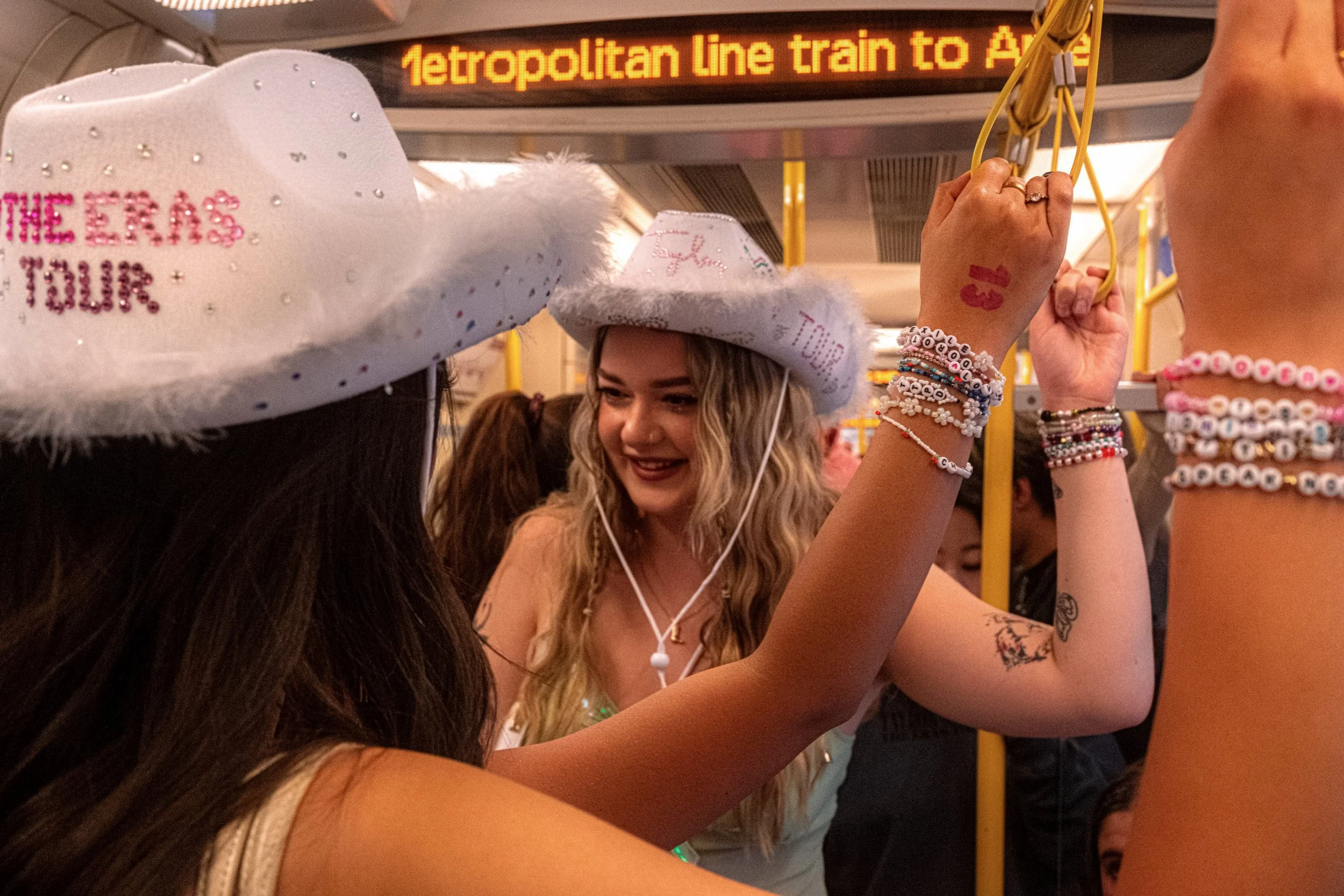 Taylor Swift fans in white cowboy hats hold overhead rails on a Metropolitan line train, displaying friendship bracelets.