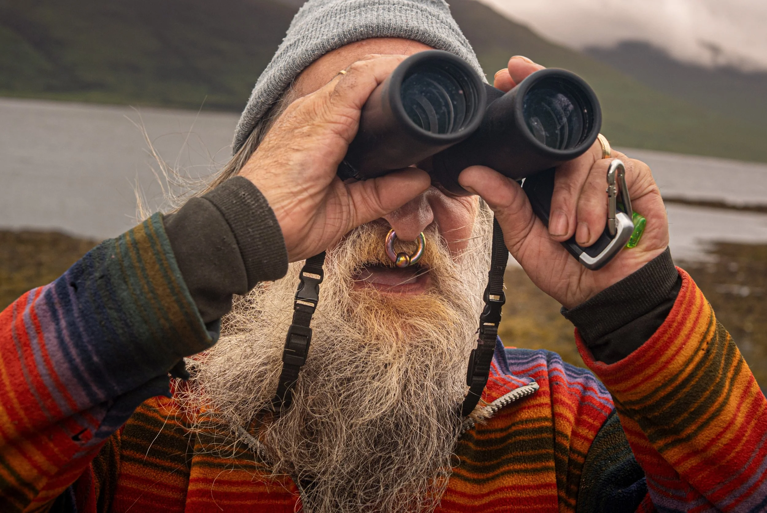 A bearded man in a striped jumper scans the hills through binoculars on the Isle of Mull.