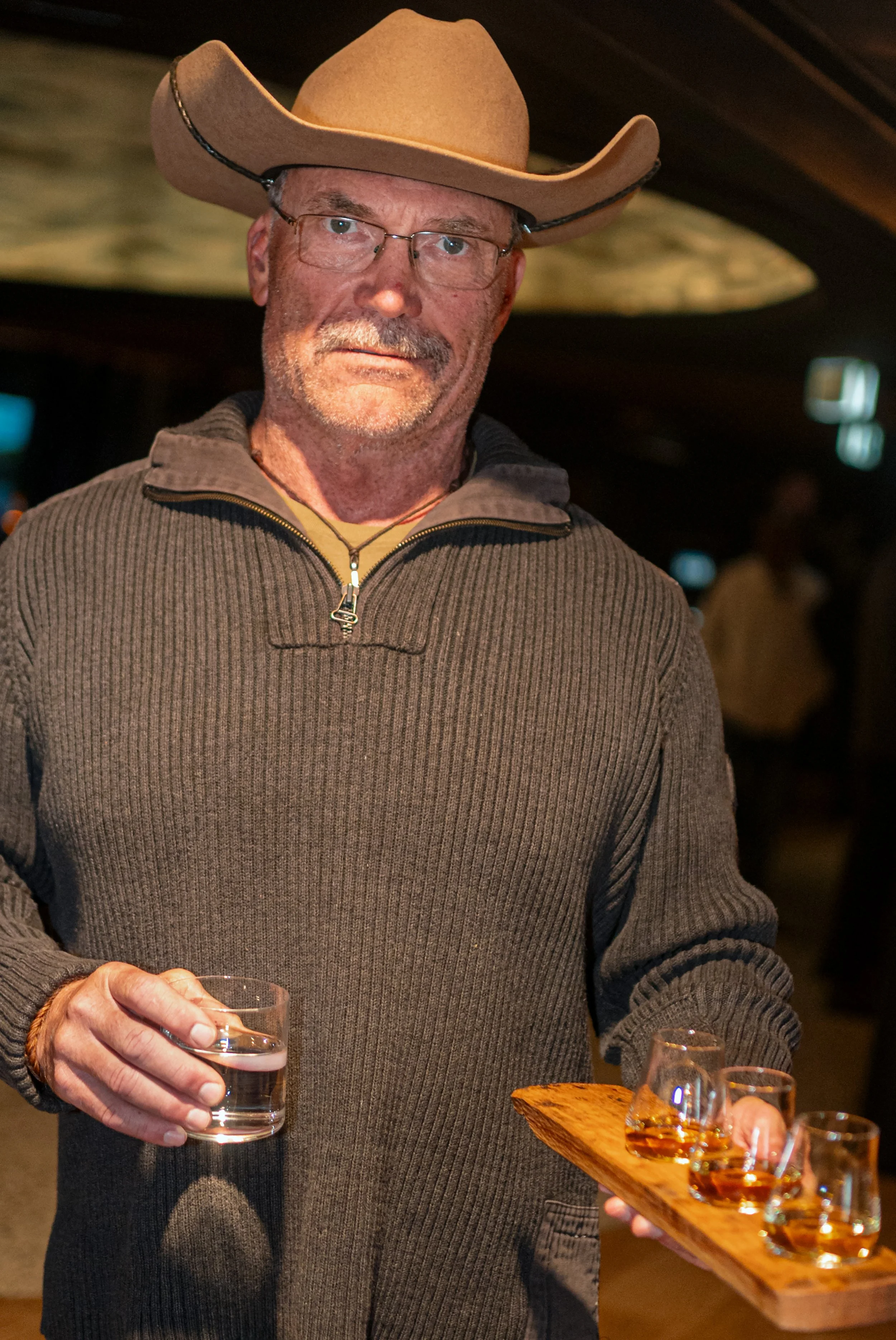 A man in a cowboy hat holds a tray of whisky samples in a dimly lit bar.