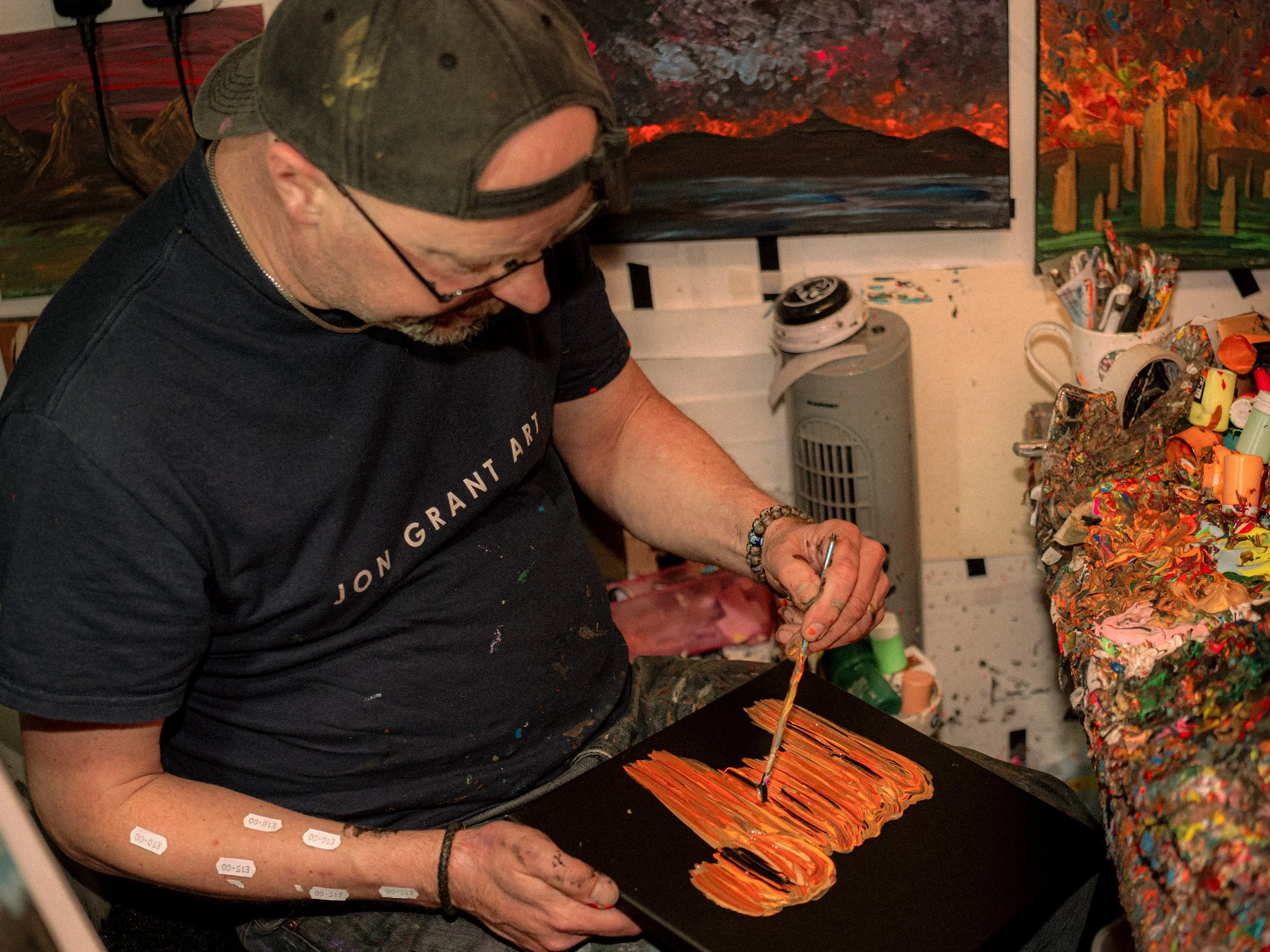 An artist in a cap paints bright orange strokes in his crowded studio.