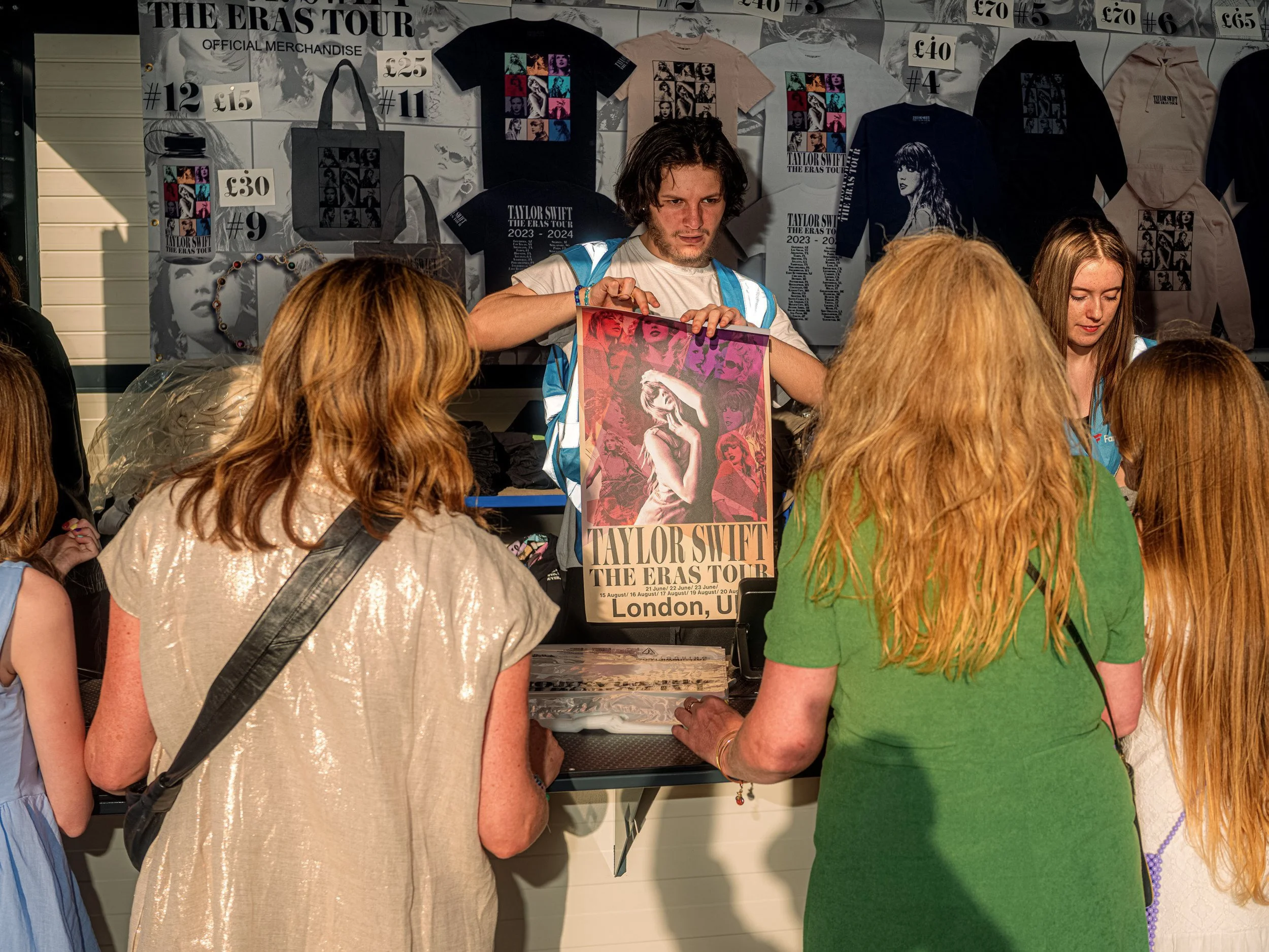 Fans browse Taylor Swift T-shirts while a stallholder displays an Eras Tour London poster at a merchandise stand.