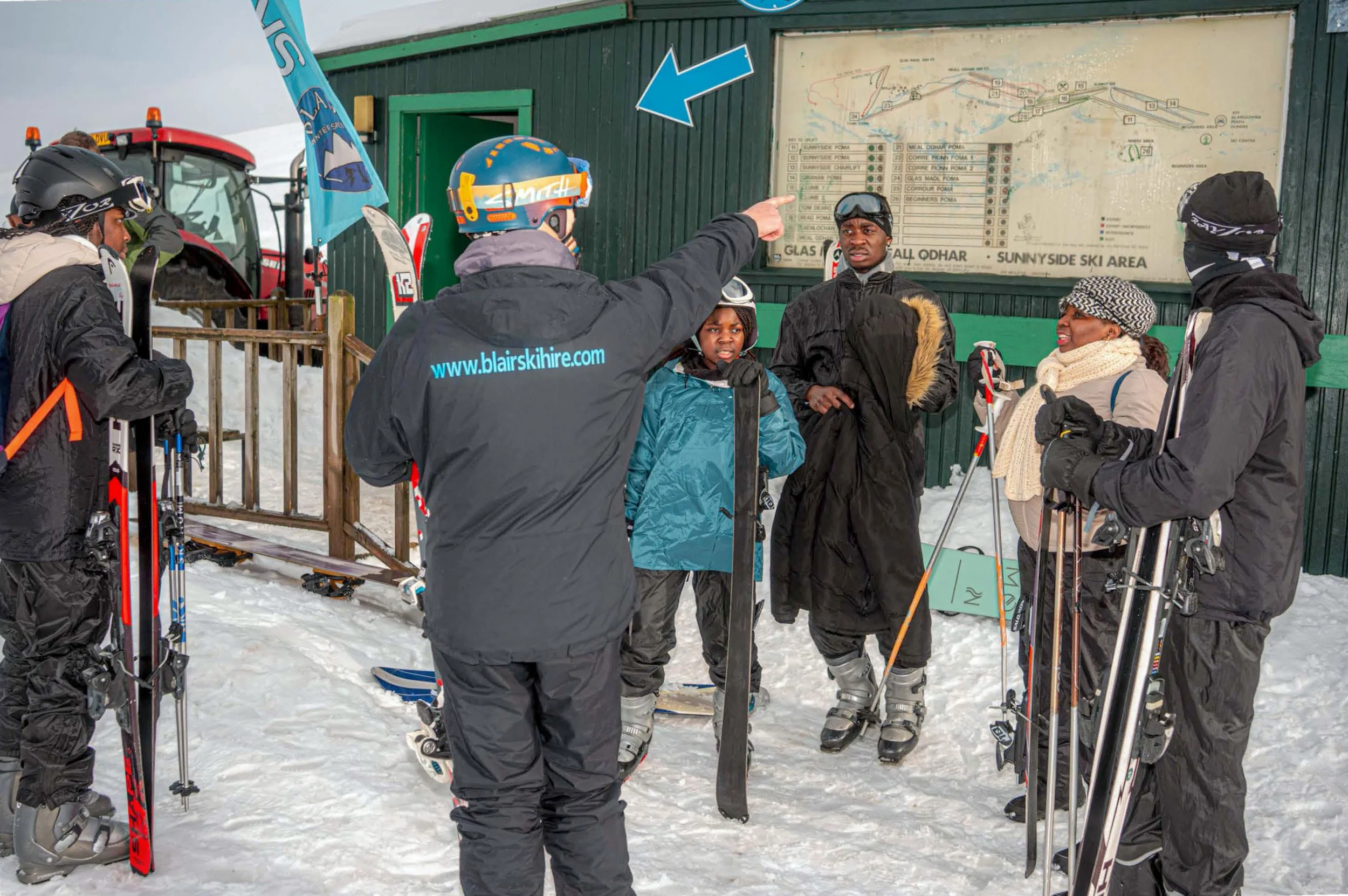 A man points at a piste map as a group of skiers gathers beside a green hut.