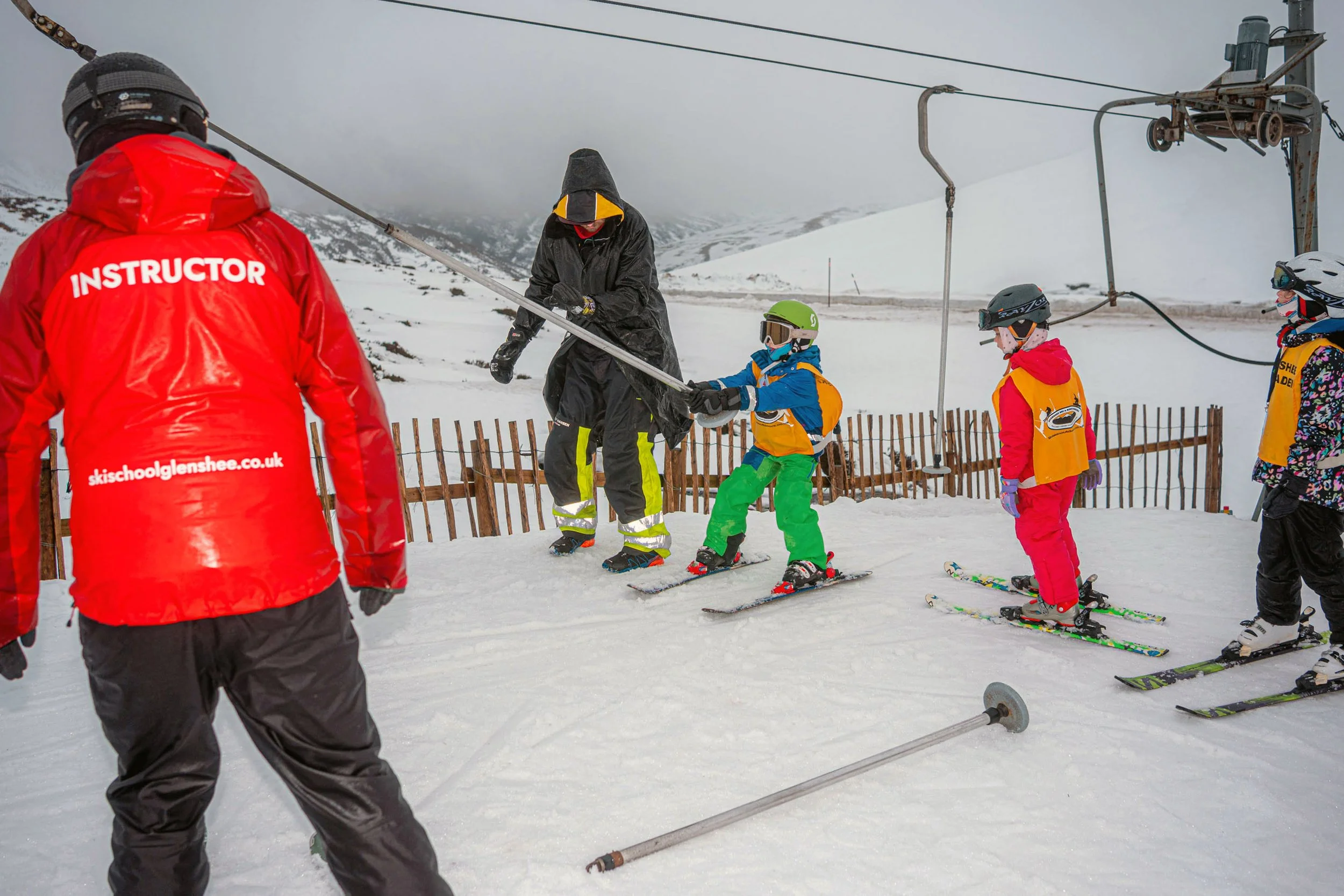 A ski instructor in a red jacket watches young children learning on a lift in the snow.