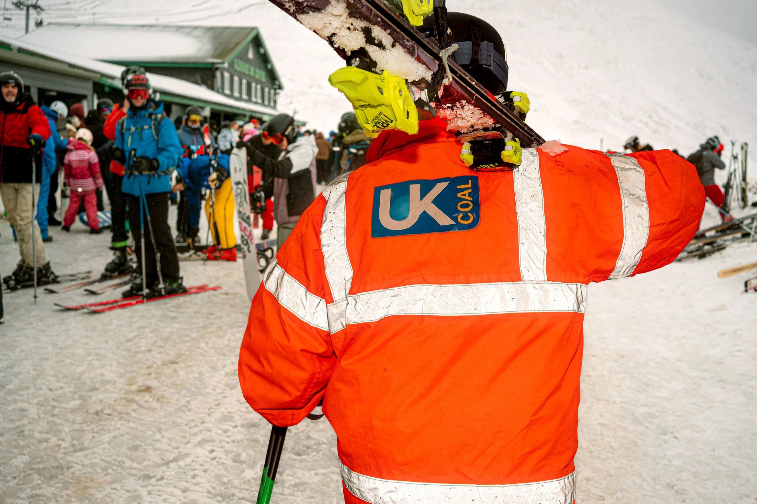 A man in an orange UK Coal hi-vis jacket carries skis on his shoulder through a crowd of skiers.