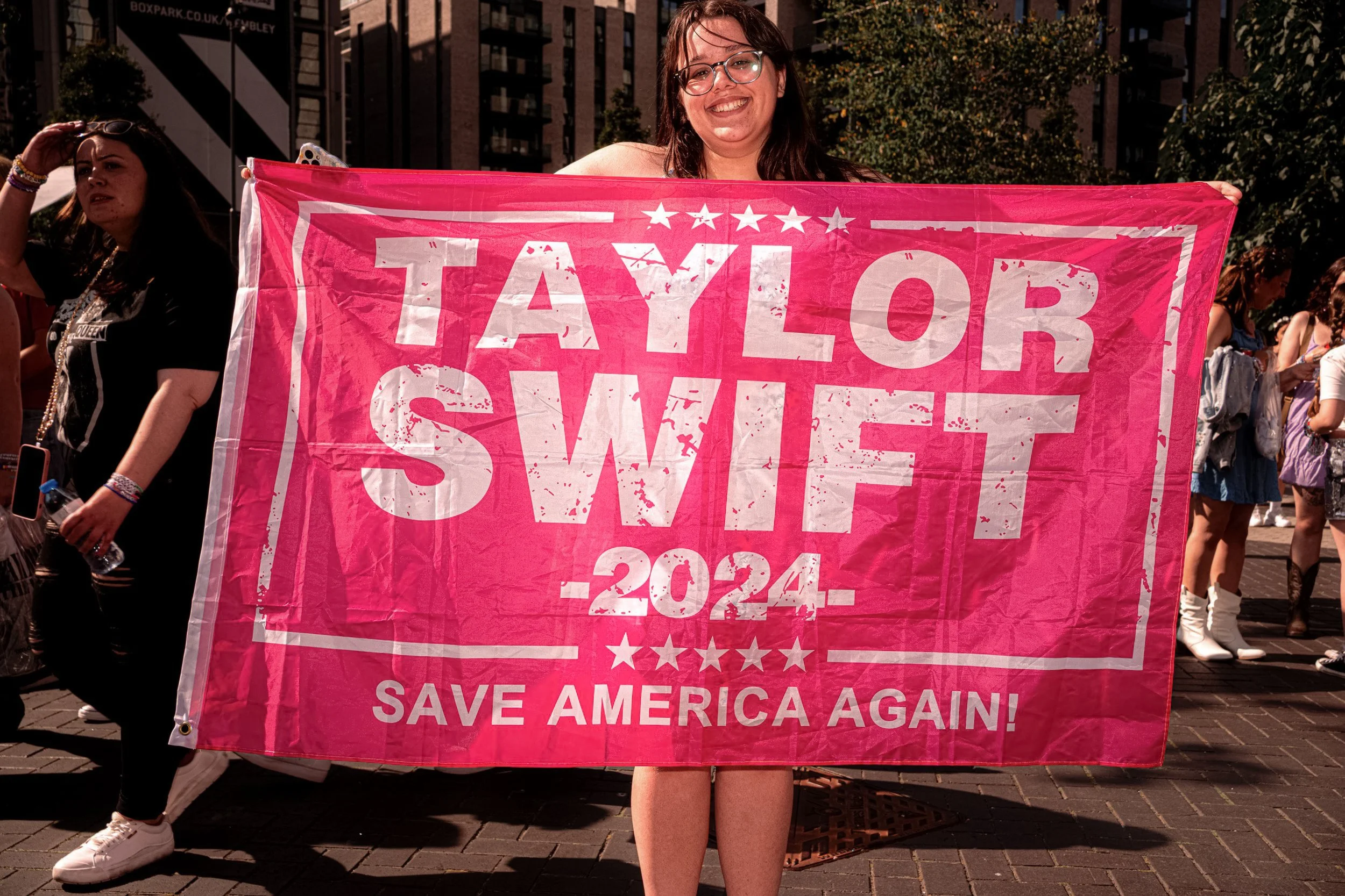 A Taylor Swift fan holds a large pink banner reading “Taylor Swift 2024 — Save America Again”.