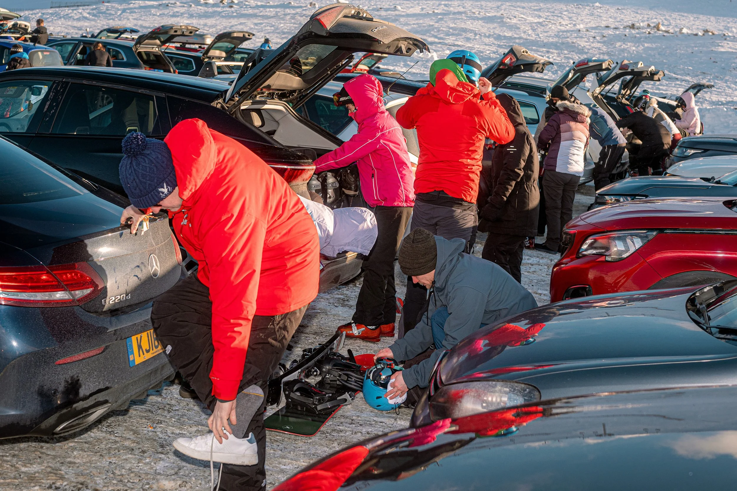 Skiers pull on boots and gear from open car boots in a packed ski centre car park.