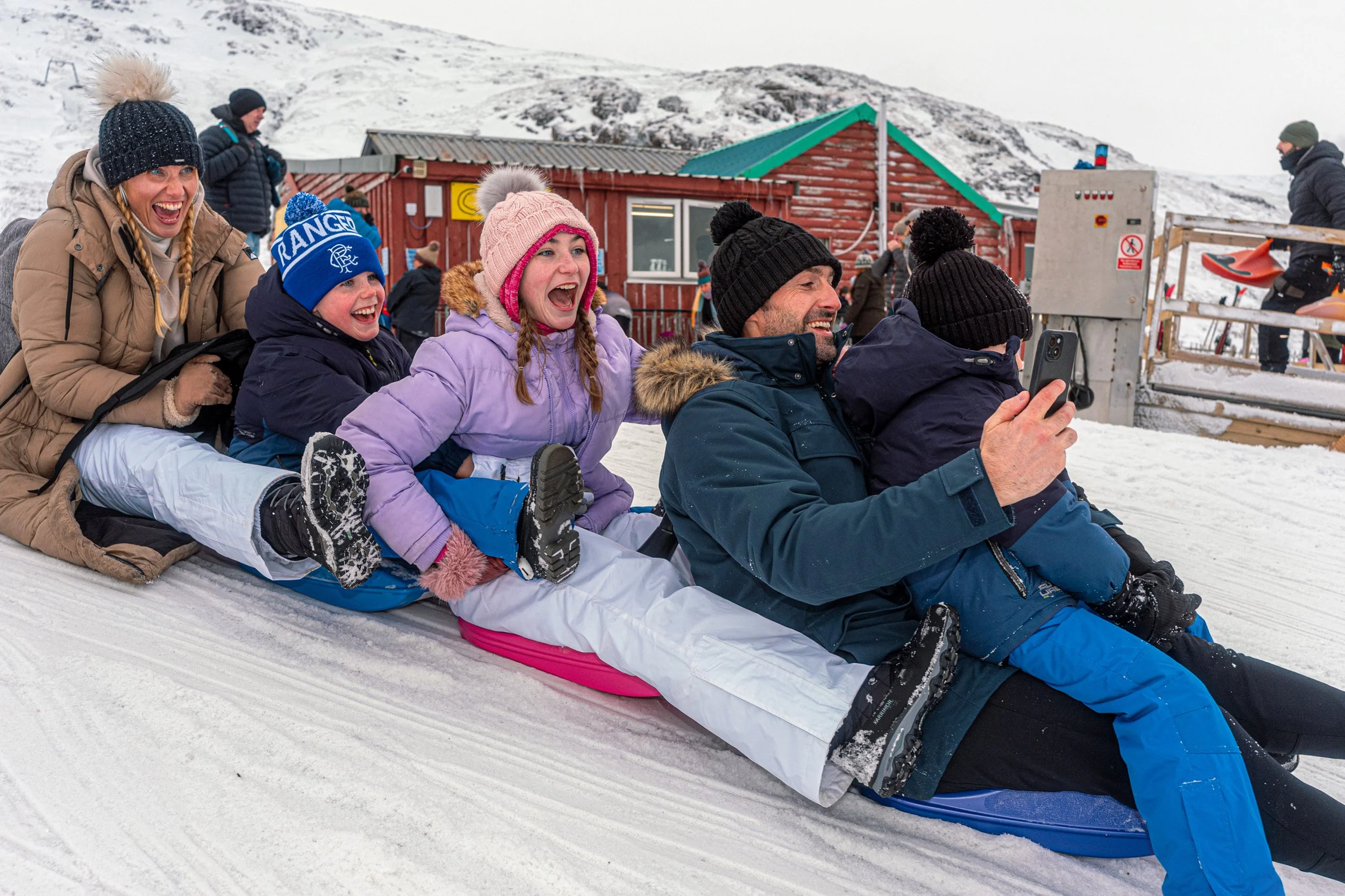 A family laughs as they slide down the snow together on sledges, one filming on a phone.