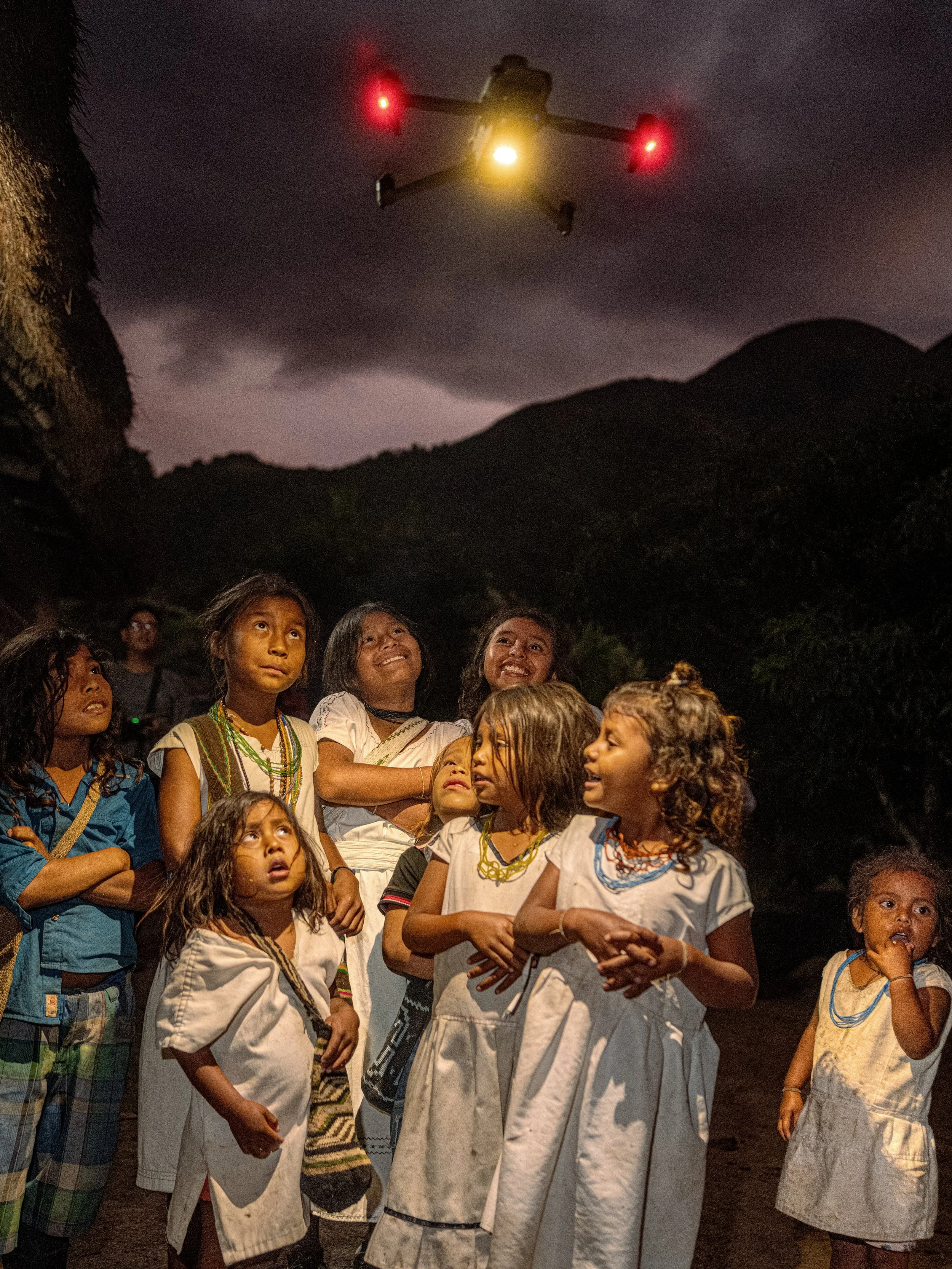 A group gathers beneath a hovering drone with red and yellow lights against a dark mountain sky.