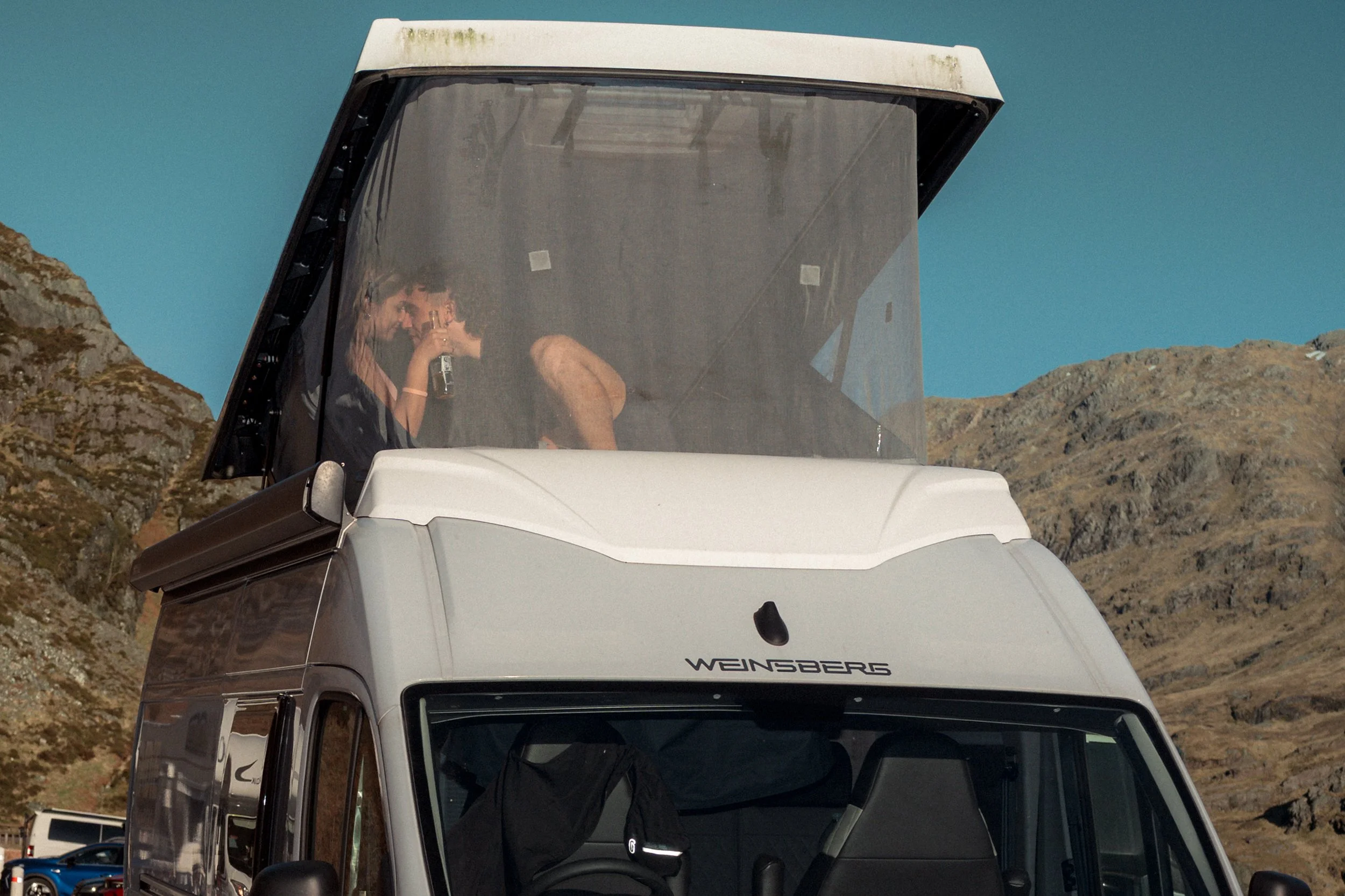 A couple sit inside the pop-up roof of a camper van against rocky hills.