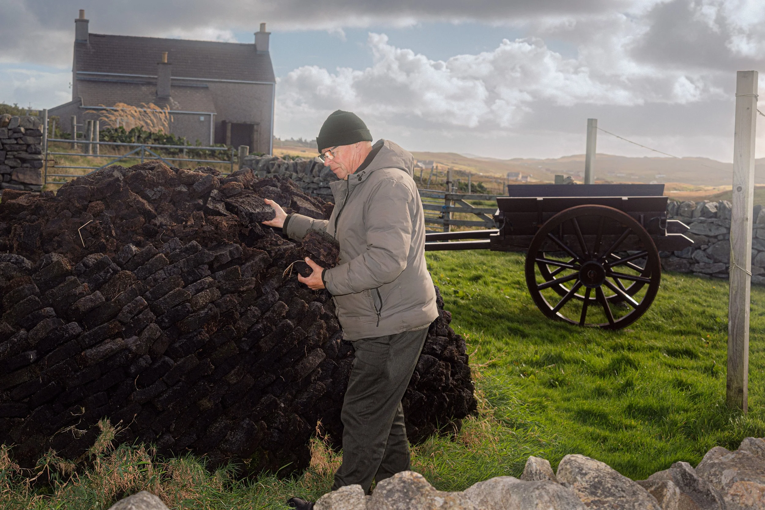 A man photographs a peat stack beside an old cart and a croft house.