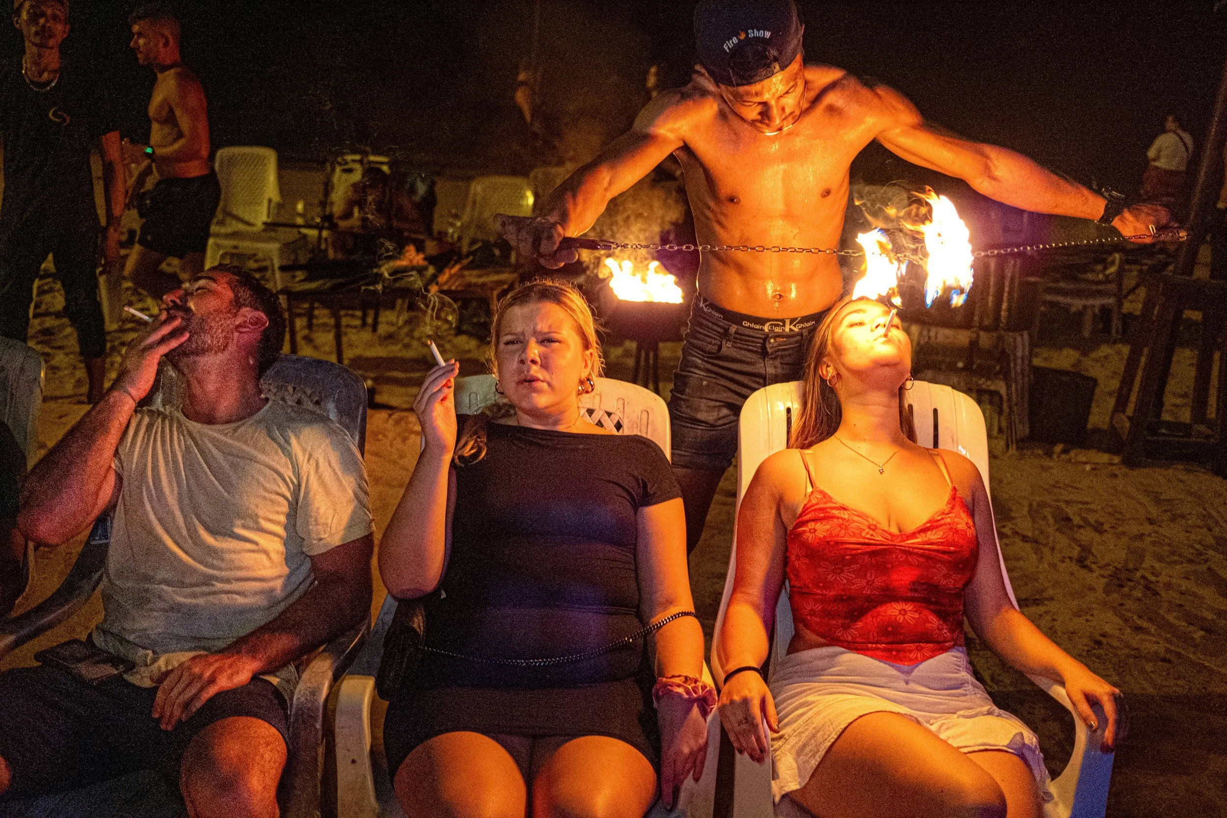 A fire performer stands behind three seated holidaymakers during a night-time show in Thailand.