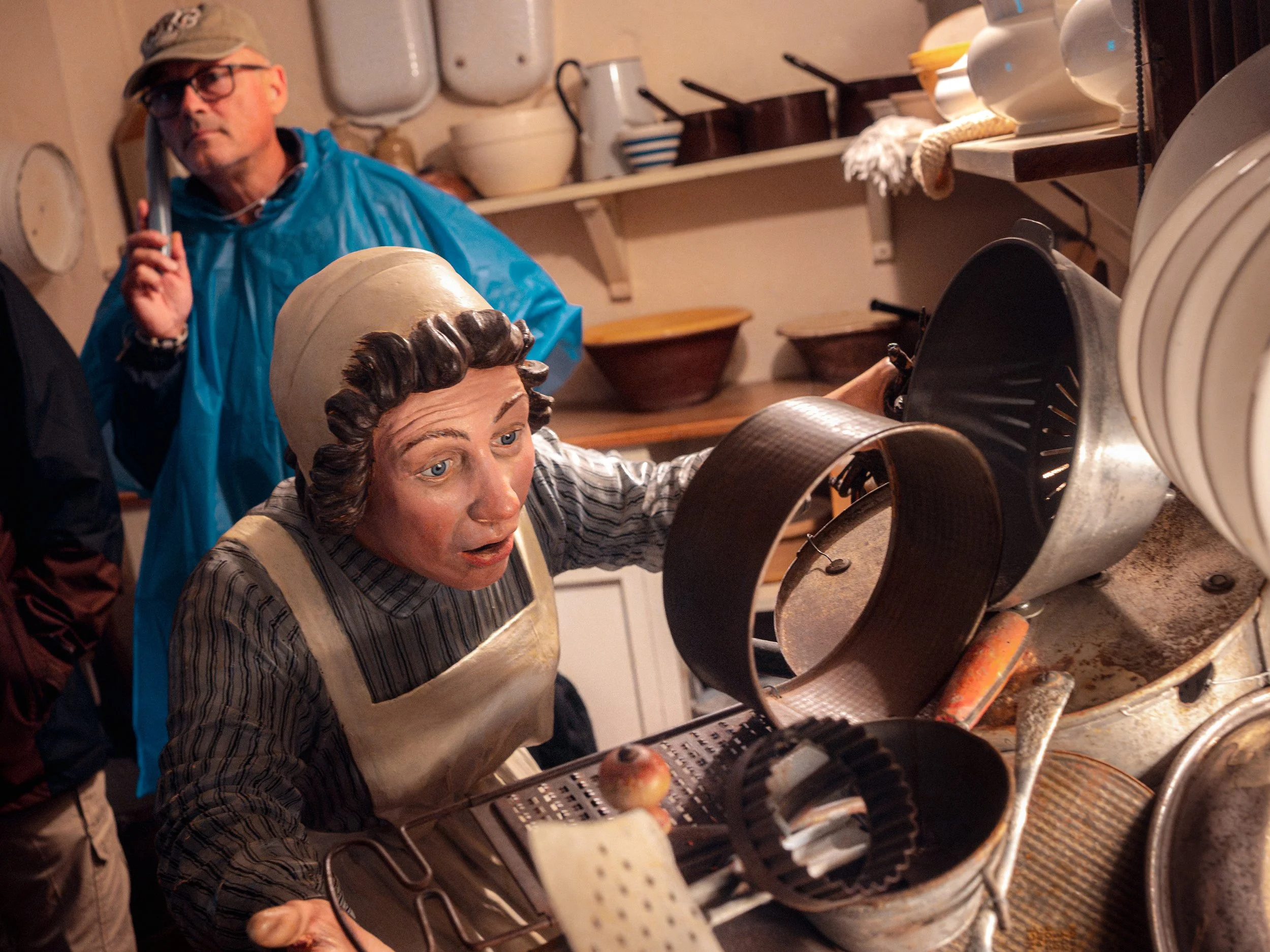 A visitor in a blue rain poncho looks at a life-size figure of a cook among pots and pans in a historic kitchen.
