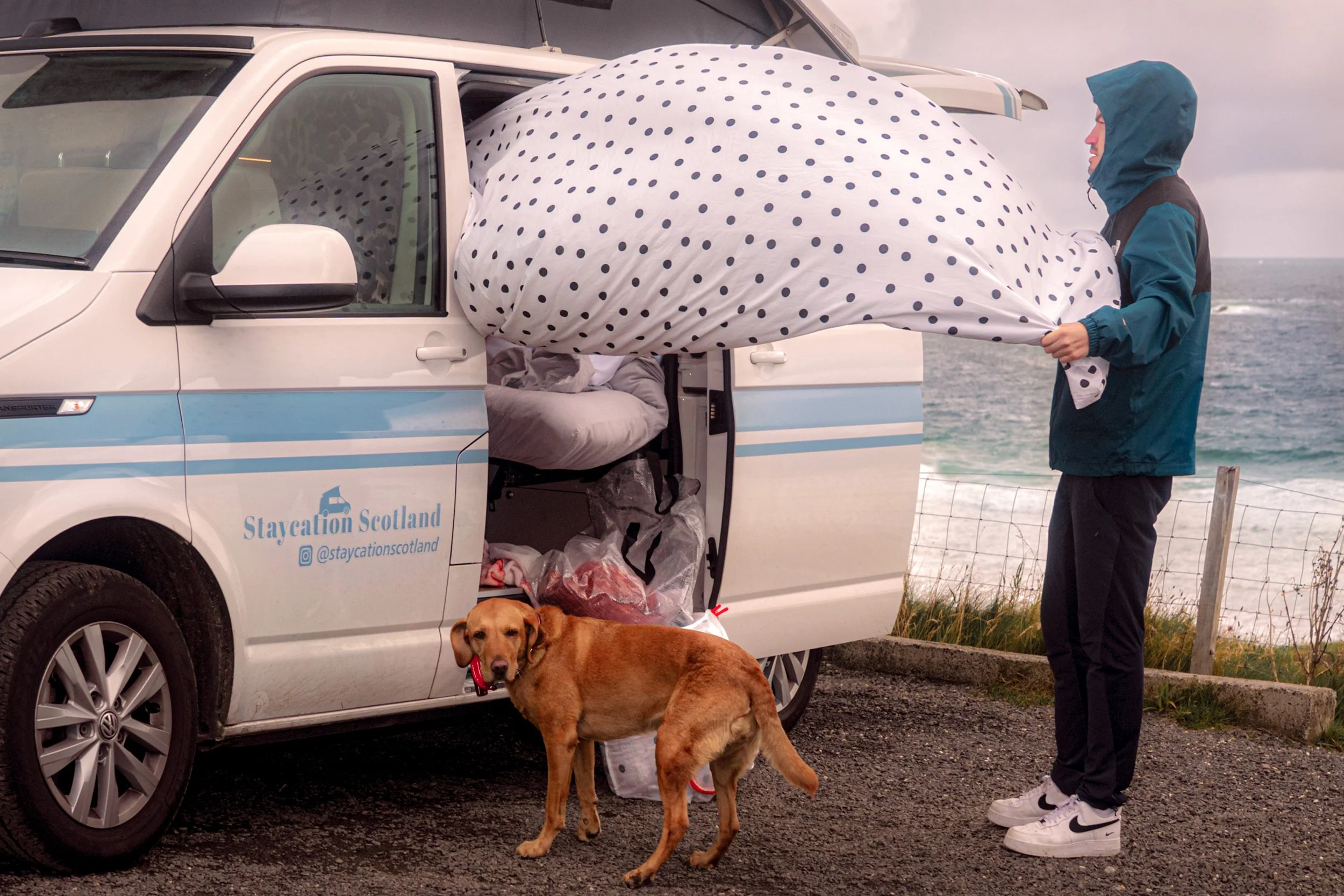 A woman holds a spotted umbrella by the open door of a Staycation Scotland camper van as her dog waits beside the sea.