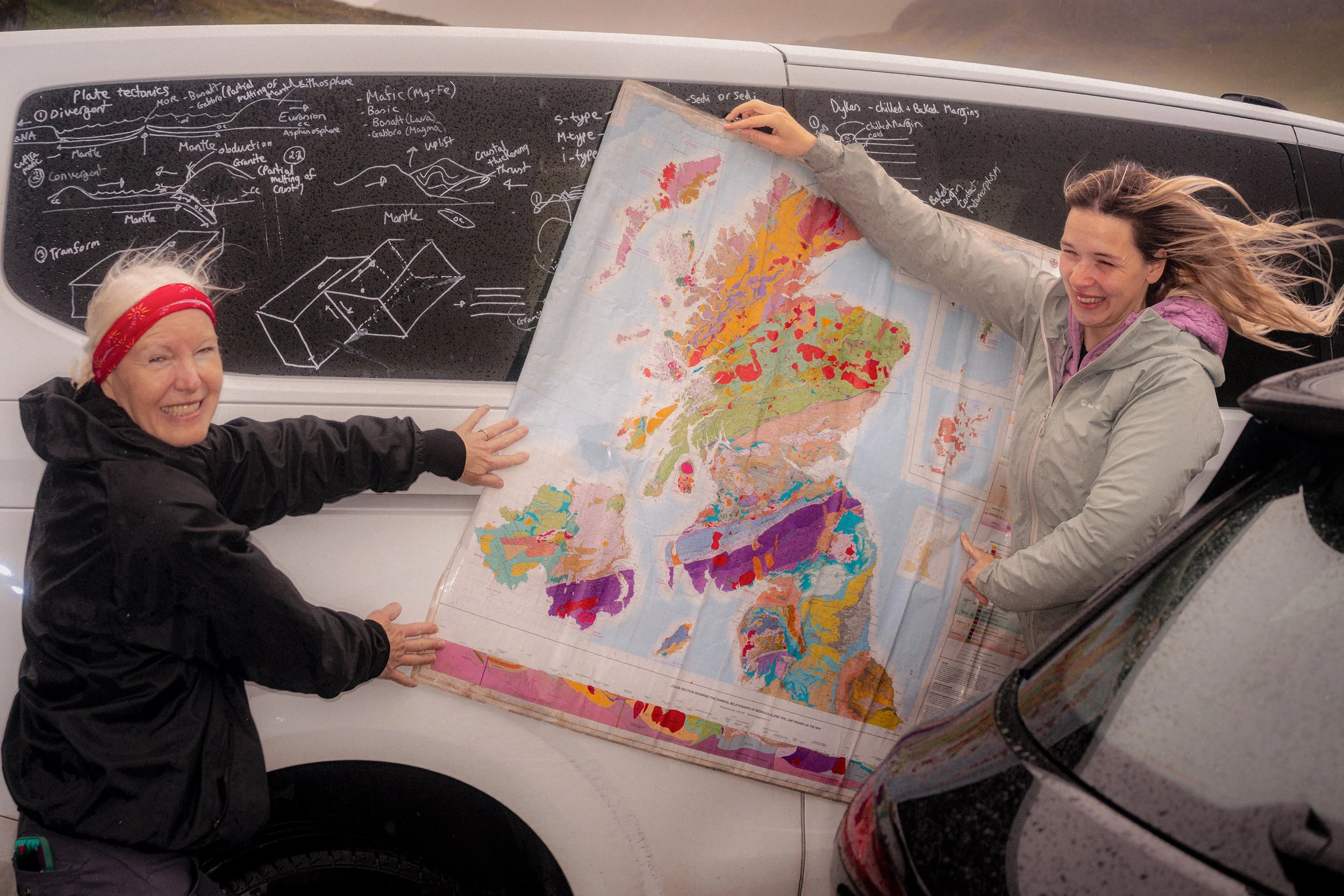 Two women hold up a geological map of Scotland against a camper van covered in chalk notes.