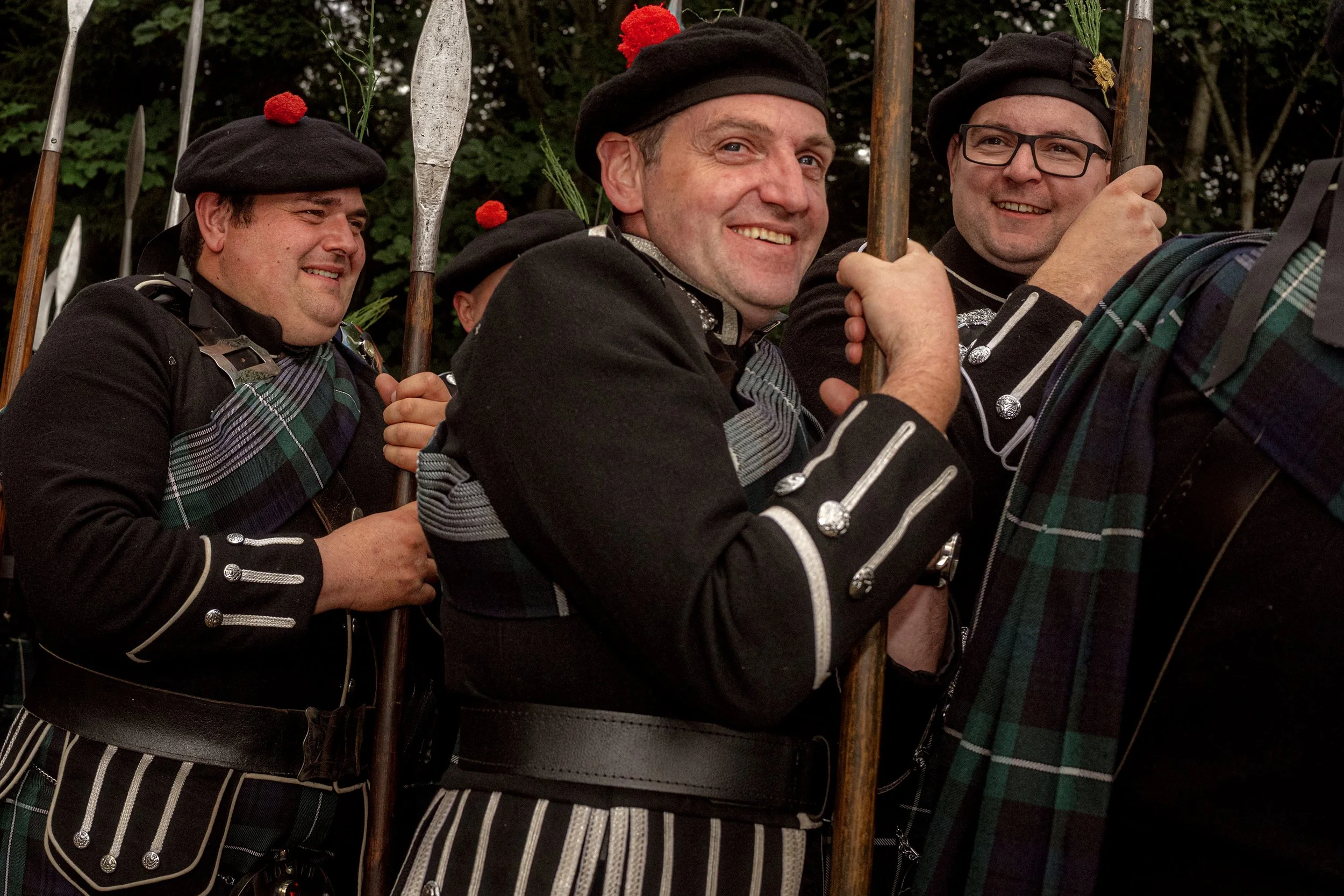 Three men in Highland dress hold pikes and smile at a Highland gathering.